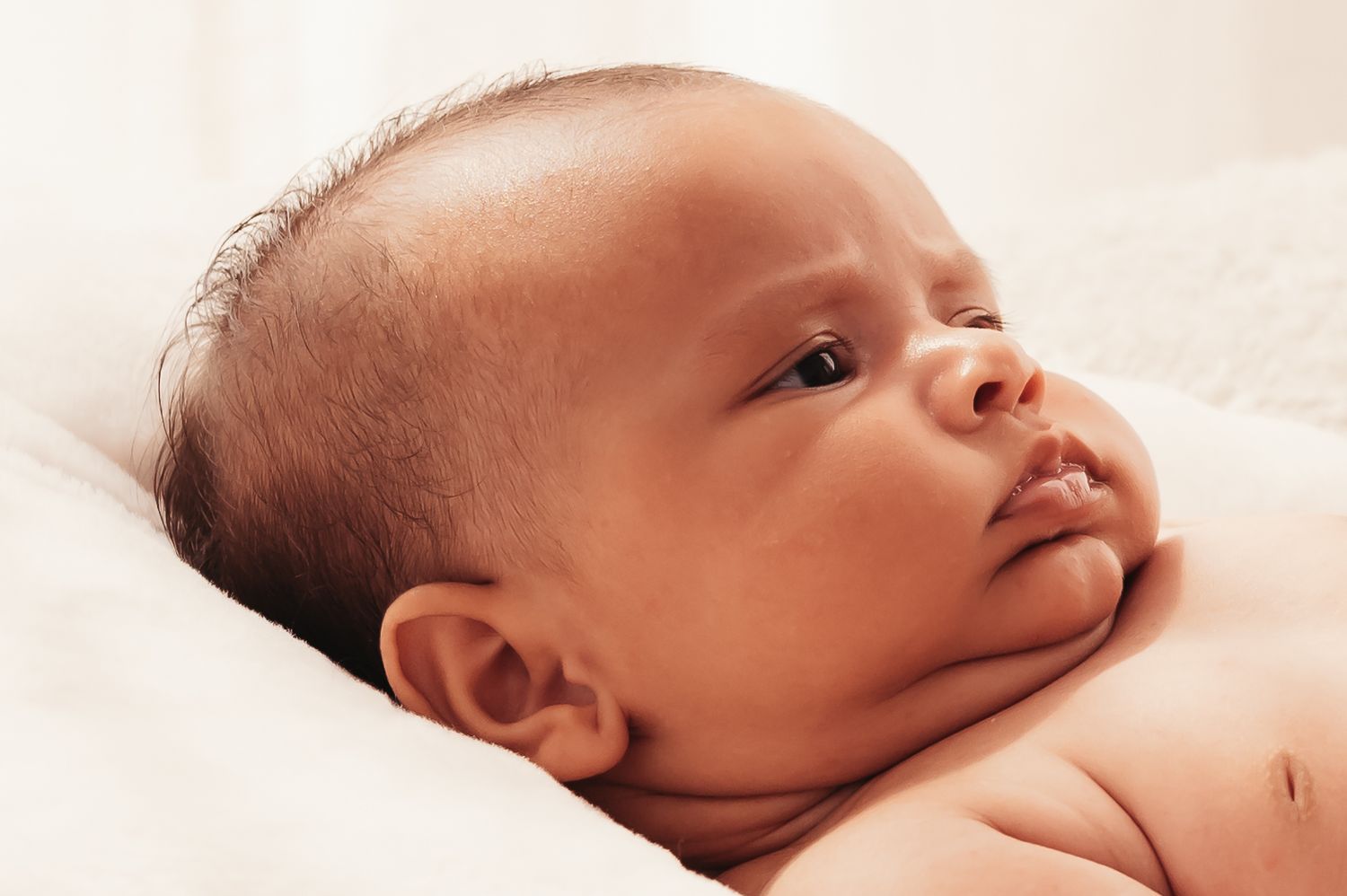 Newborn baby resting on white bedding with eyes gazing upward in gentle contemplation.