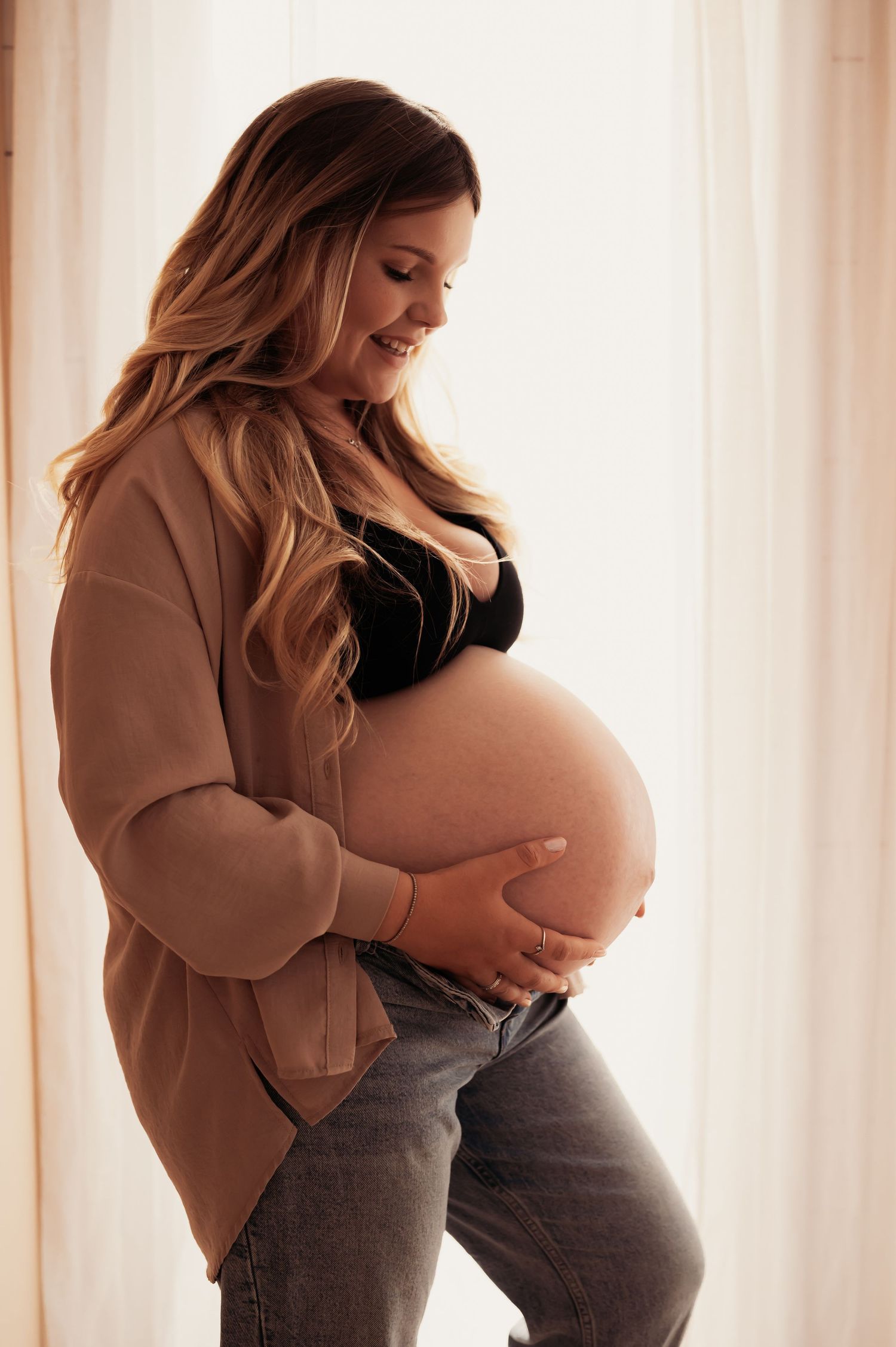 Smiling pregnant woman in black bralette, tan cardigan, and jeans cradling her baby bump near a bright window.