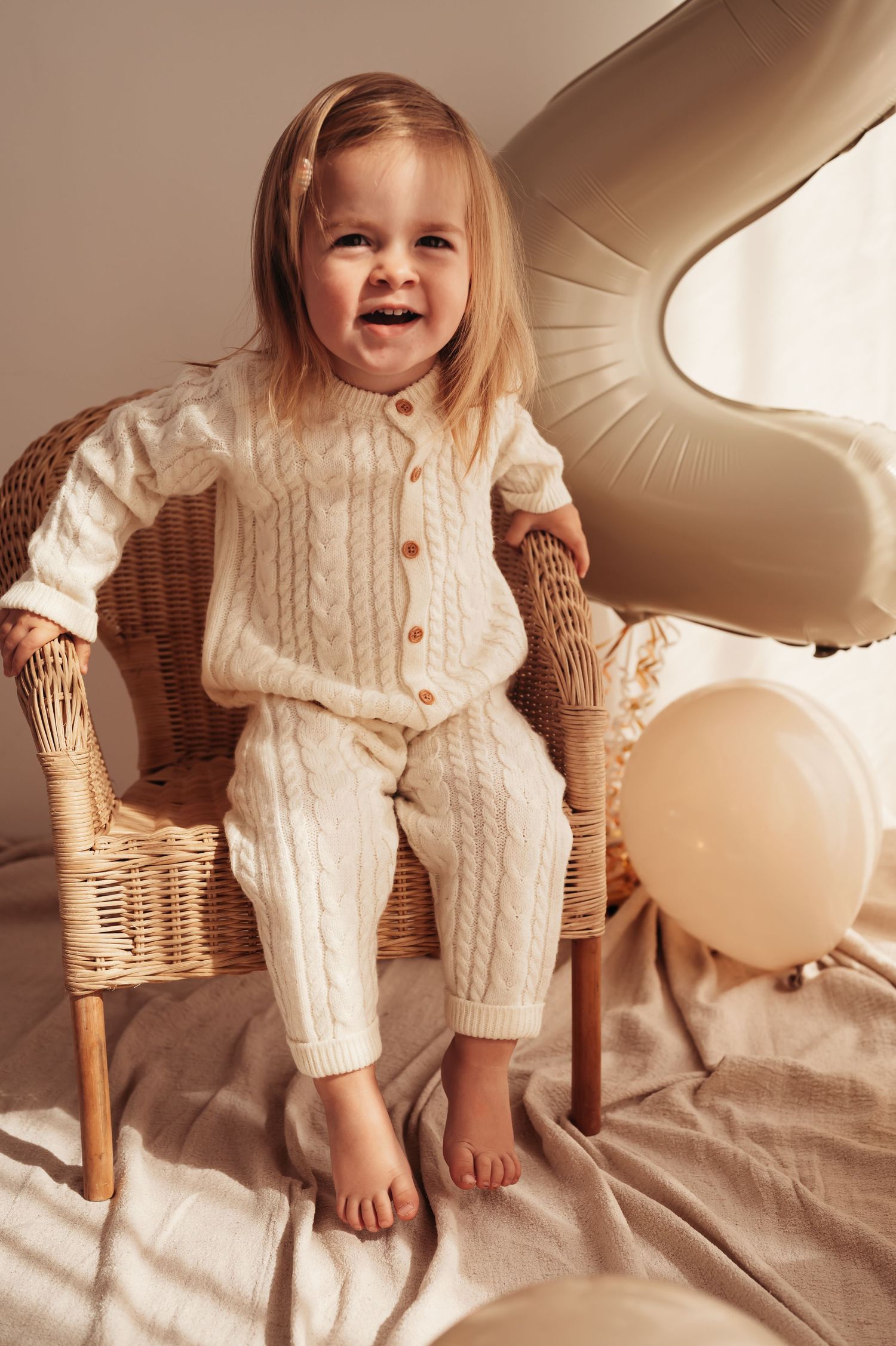 Child wearing cozy cream knit outfit sits in wicker chair with balloons in warm neutral setting.