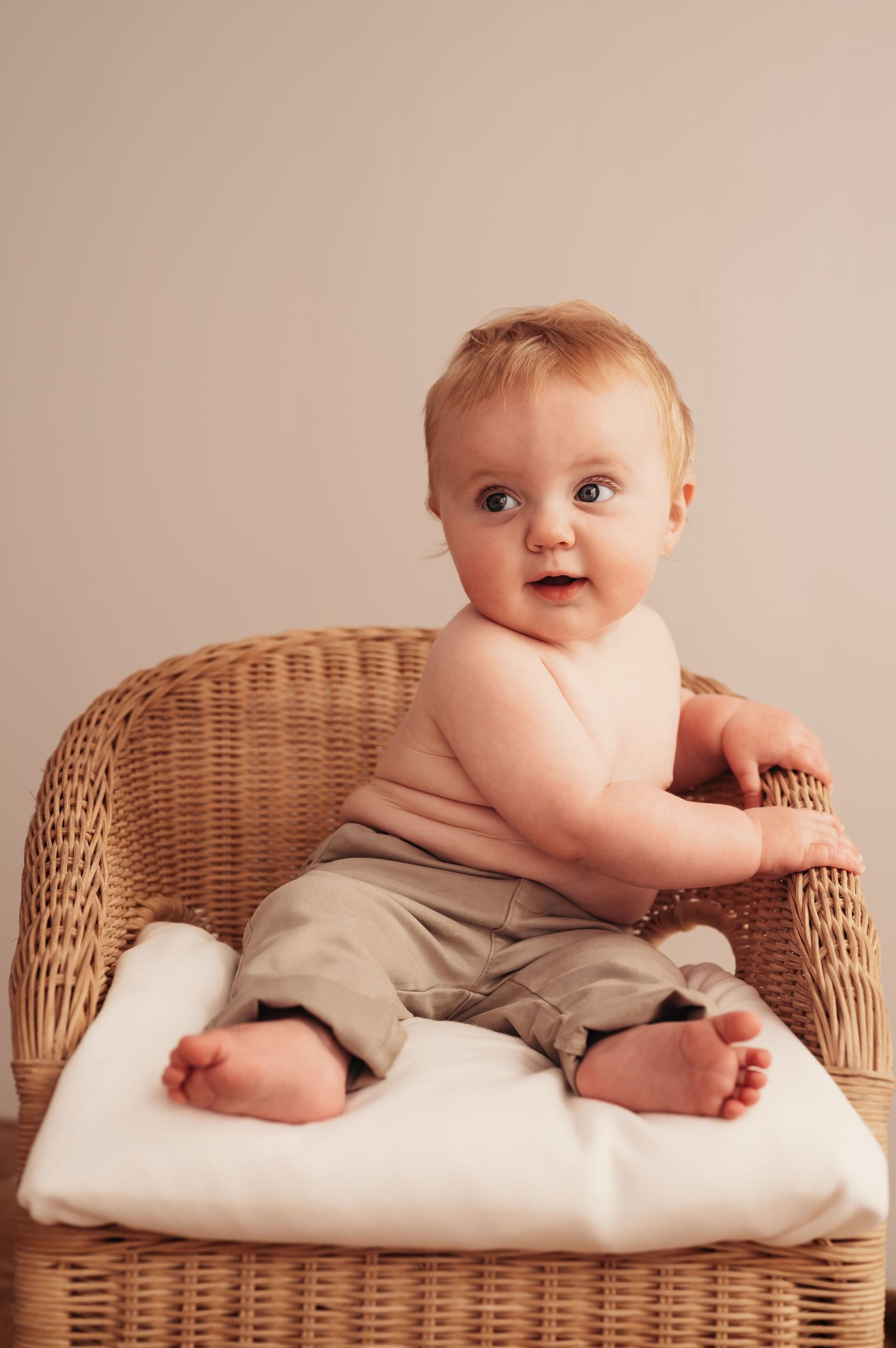 Baby sits in wicker chair on white cushion wearing khaki pants.