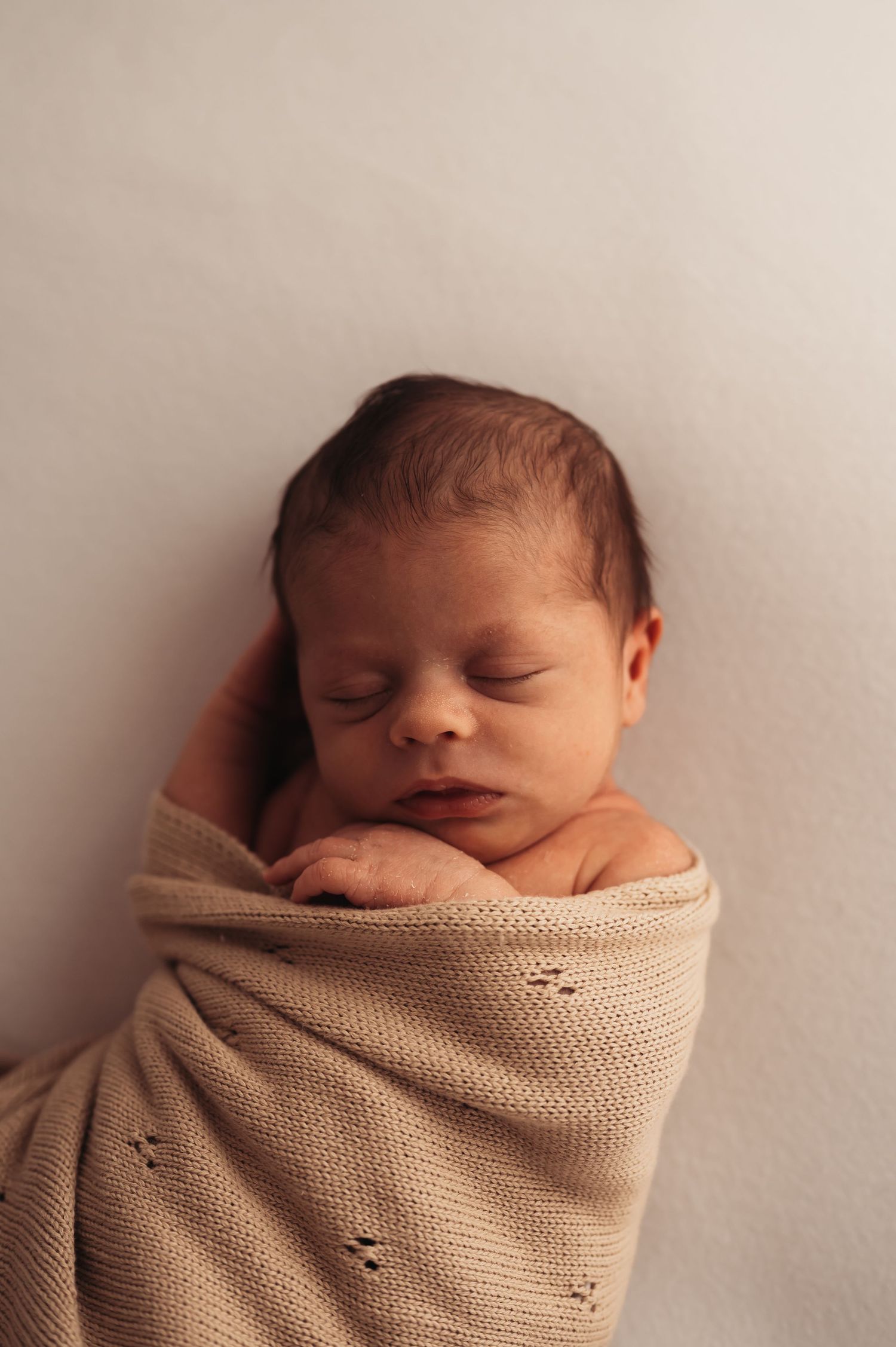 Newborn baby swaddled in beige knitted wrap against neutral background during portrait session.