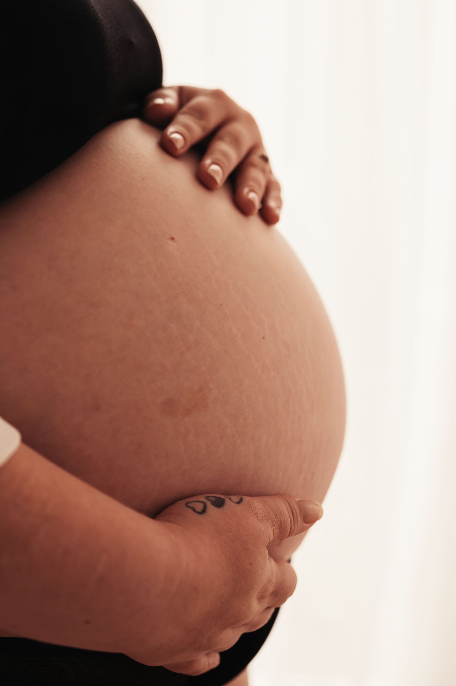 Close-up view of hands cradling a pregnant belly with small tattoo visible.