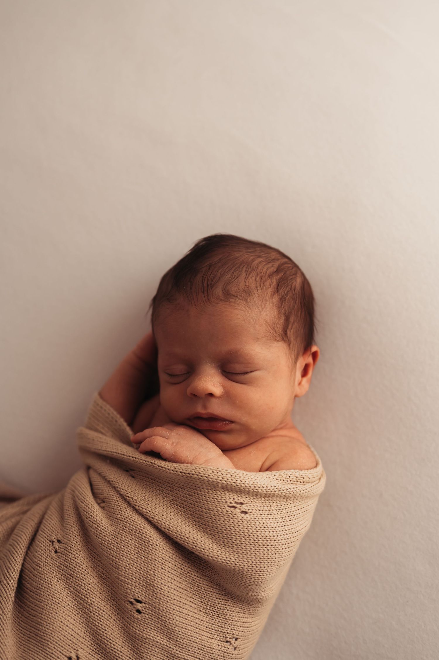 Peaceful newborn baby swaddled in a soft beige knit blanket, sleeping with hands tucked under chin against a white background.