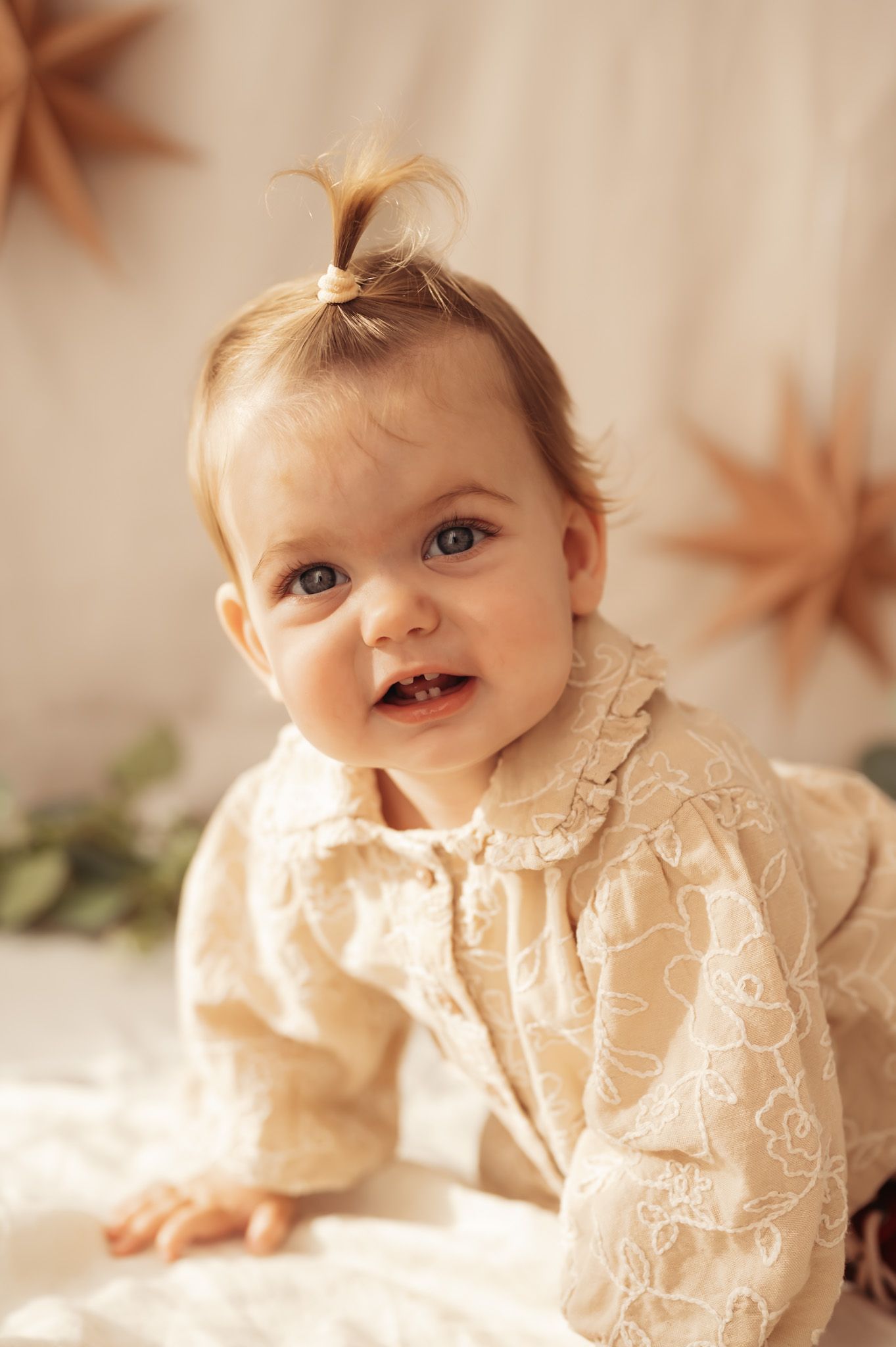 Adorable baby girl in a cream lace dress with a tiny hair bow, smiling with star decorations in background.
