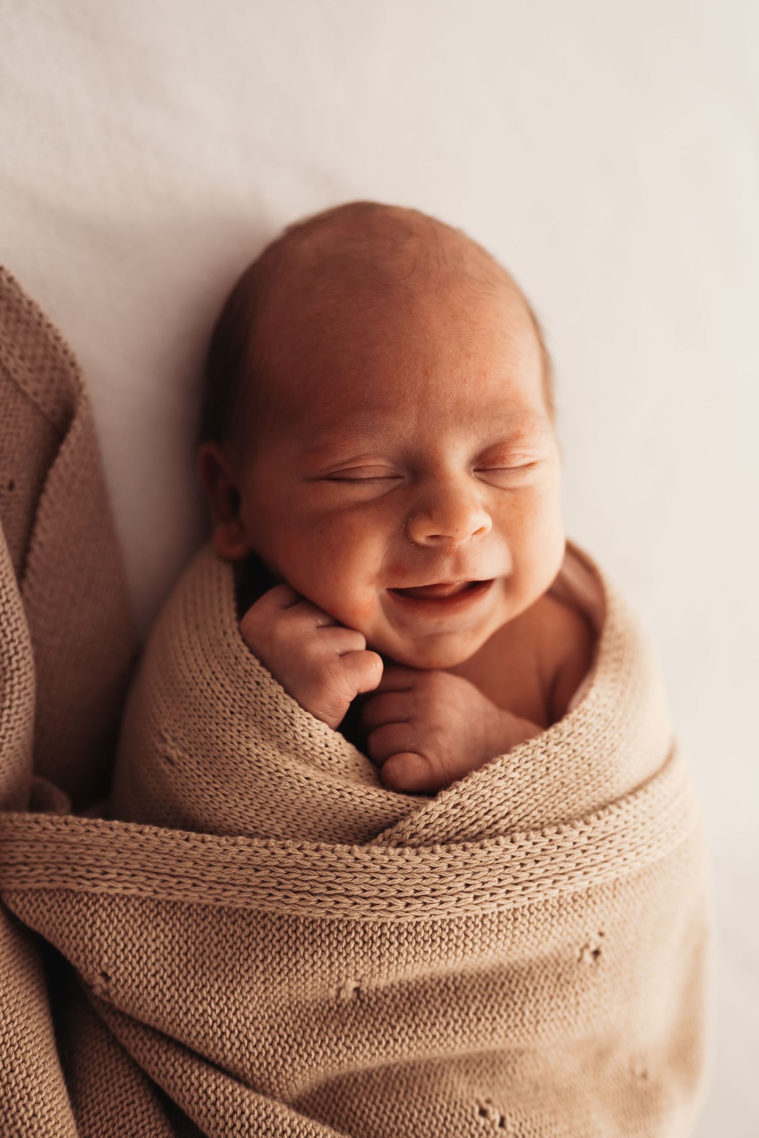 Smiling newborn baby wrapped in beige knitted blanket during photoshoot.