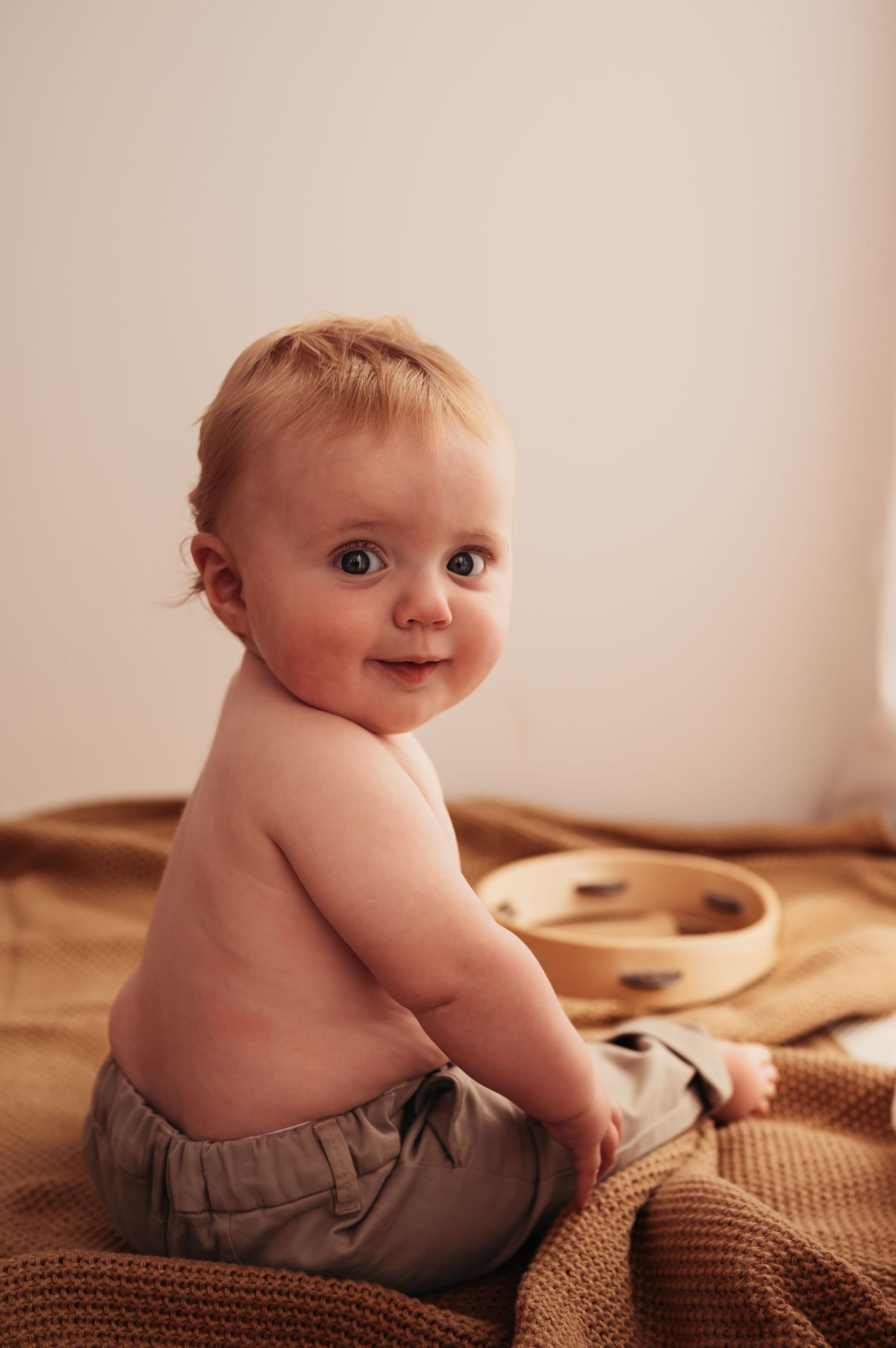 Baby in khaki pants turns to smile during photo session on brown blanket.
