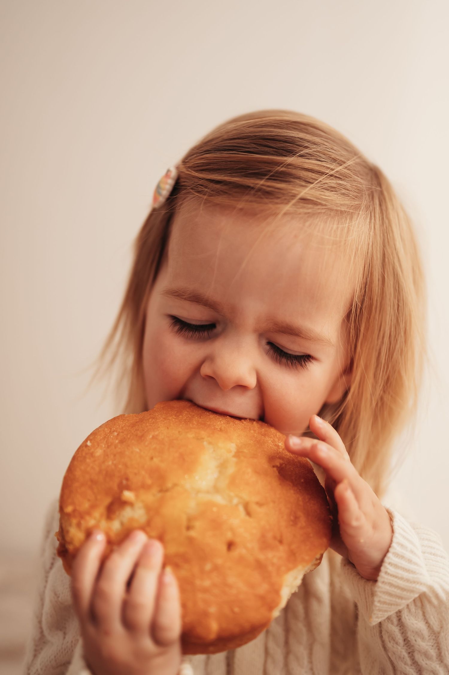 A young child in white sweater taking a big bite of cake.