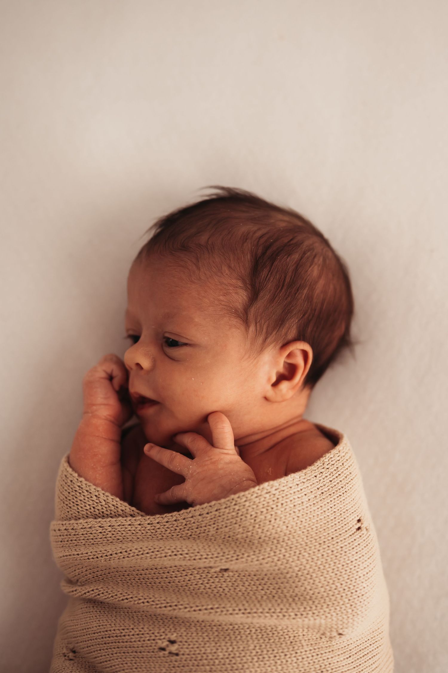 Newborn baby wrapped in beige knit blanket touching face with tiny hands.