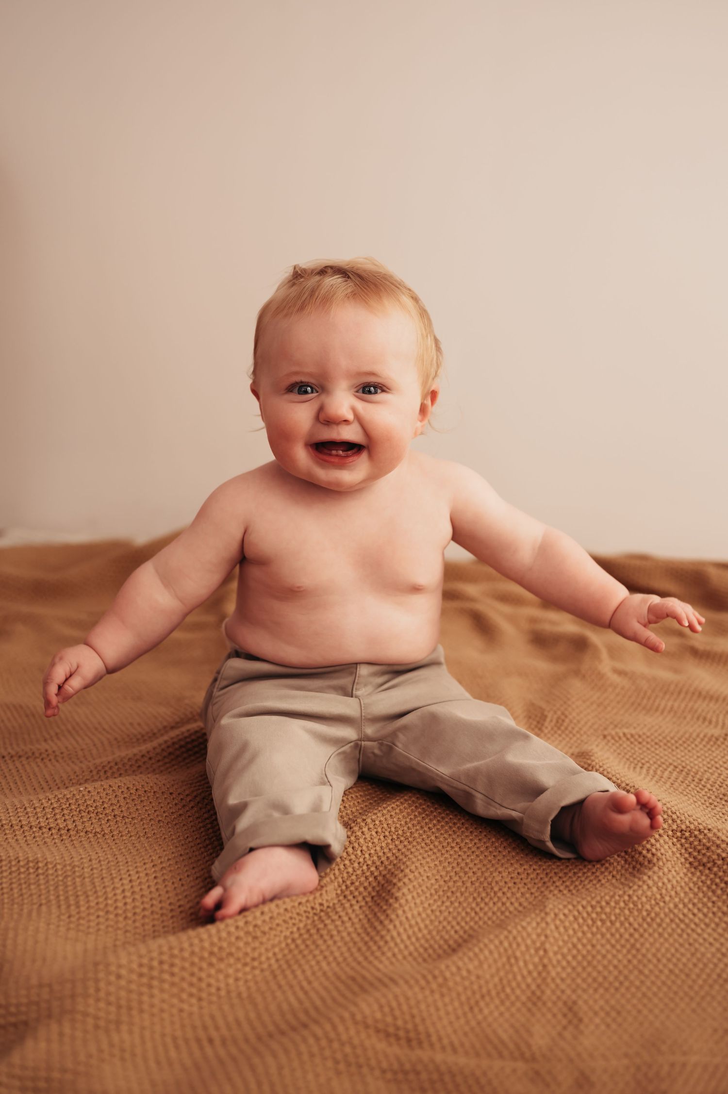 Smiling baby sits on brown textured blanket wearing khaki pants.