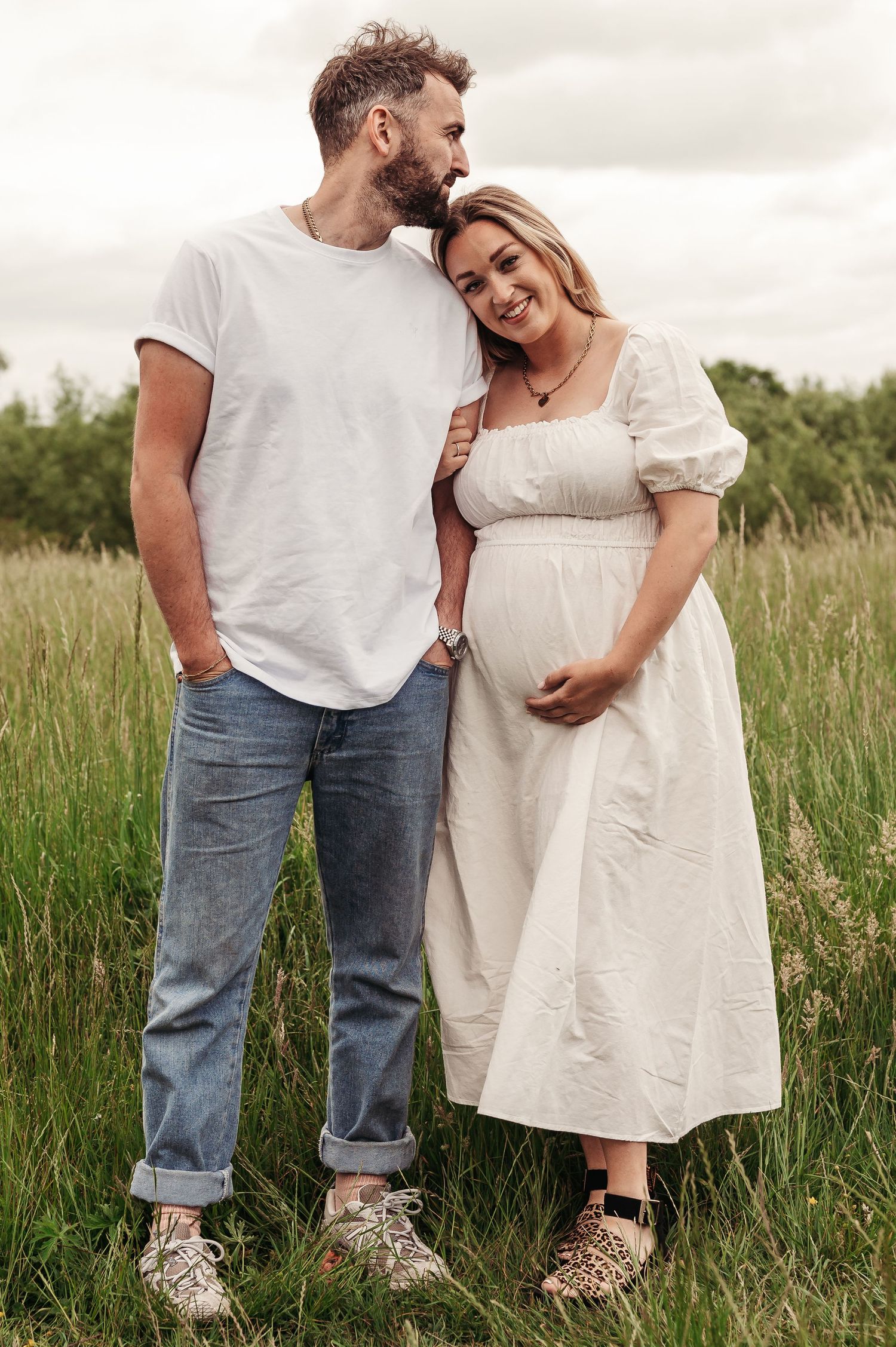 A couple stands together in a grassy field, with one wearing a white t-shirt and jeans, and the other in a flowy white dress.