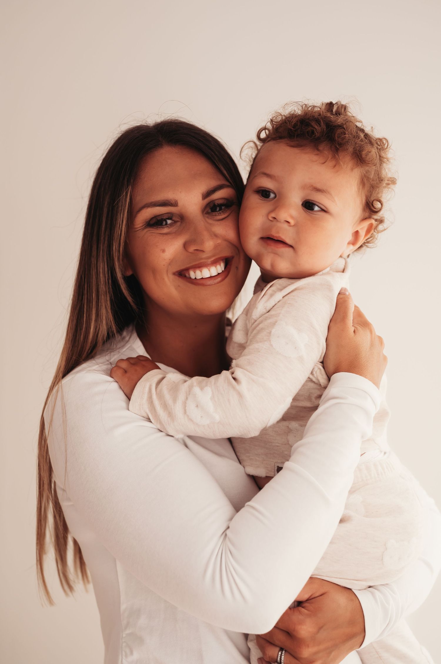 A parent in a white top holds a smiling toddler wearing a white outfit against a neutral background.