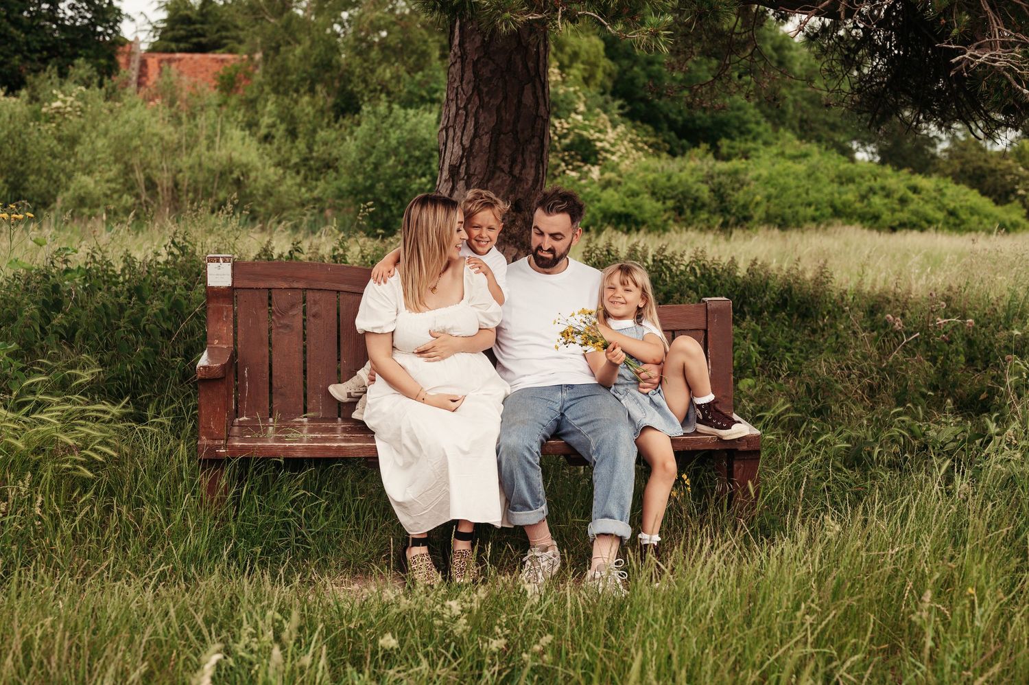 A family sits together on a wooden bench surrounded by tall grass in a natural outdoor setting.