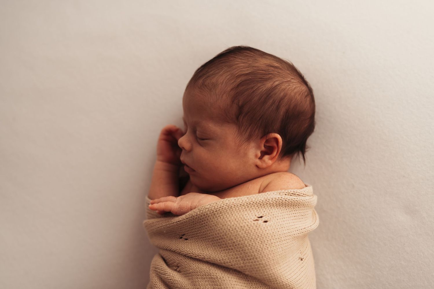 Peaceful sleeping newborn wrapped in beige knit blanket laying against soft neutral background.