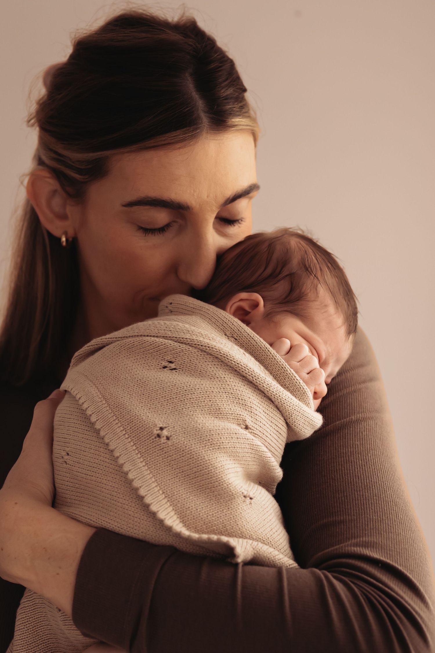 A mother tenderly kisses her sleeping newborn baby wrapped in a soft knit blanket.