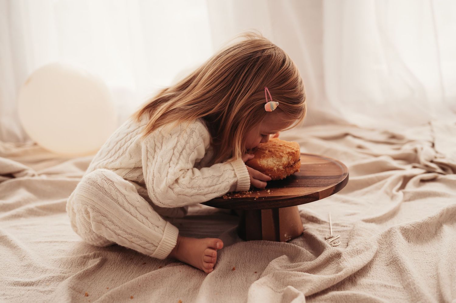 Side view of toddler in knit sweater bent over wooden cake stand on beige fabric.