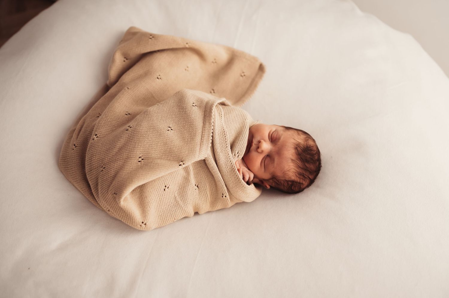 Overhead view of sleeping newborn swaddled in beige blanket on white surface.