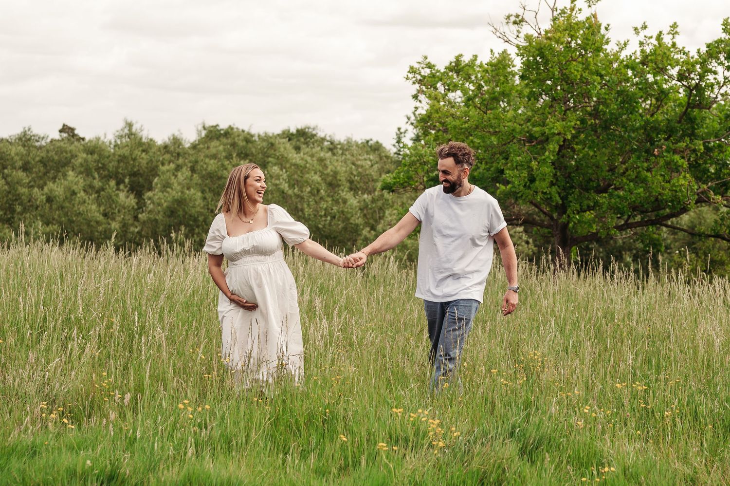 Two people walk hand in hand through a sunny meadow with tall grass and green trees in the background.