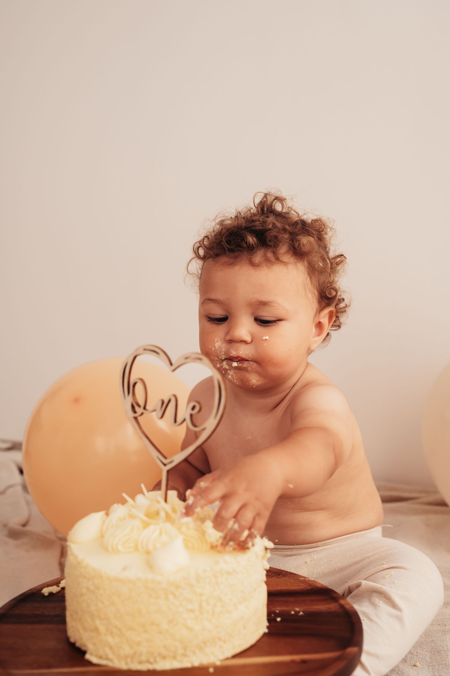 Baby reaching for white birthday cake with 'one' heart topper on wooden board.