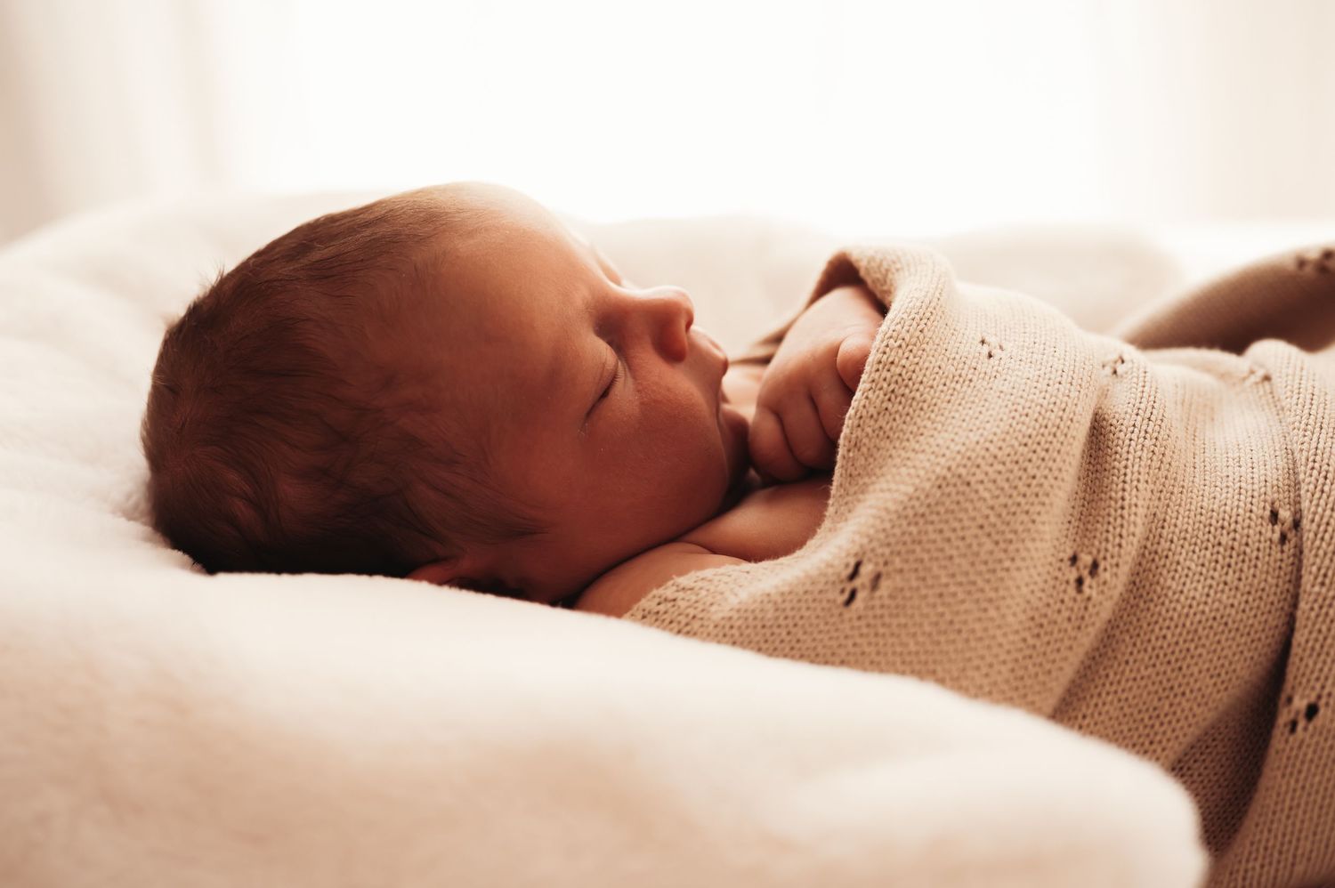 Peaceful newborn baby sleeping wrapped in a soft beige knit blanket on a white cushion.