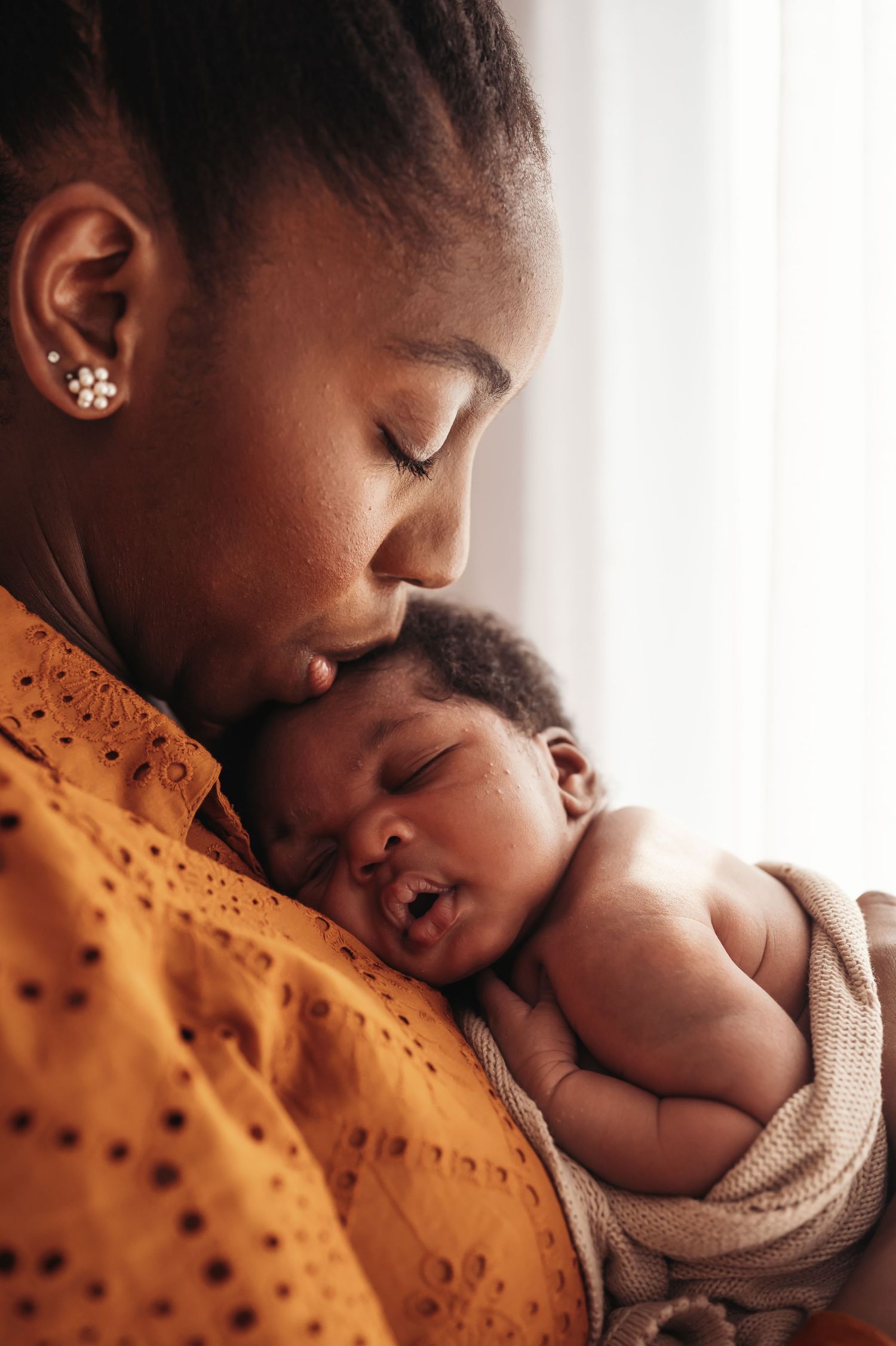 A Black mother tenderly kisses her sleeping newborn baby wrapped in a blanket, sharing a peaceful bonding moment.