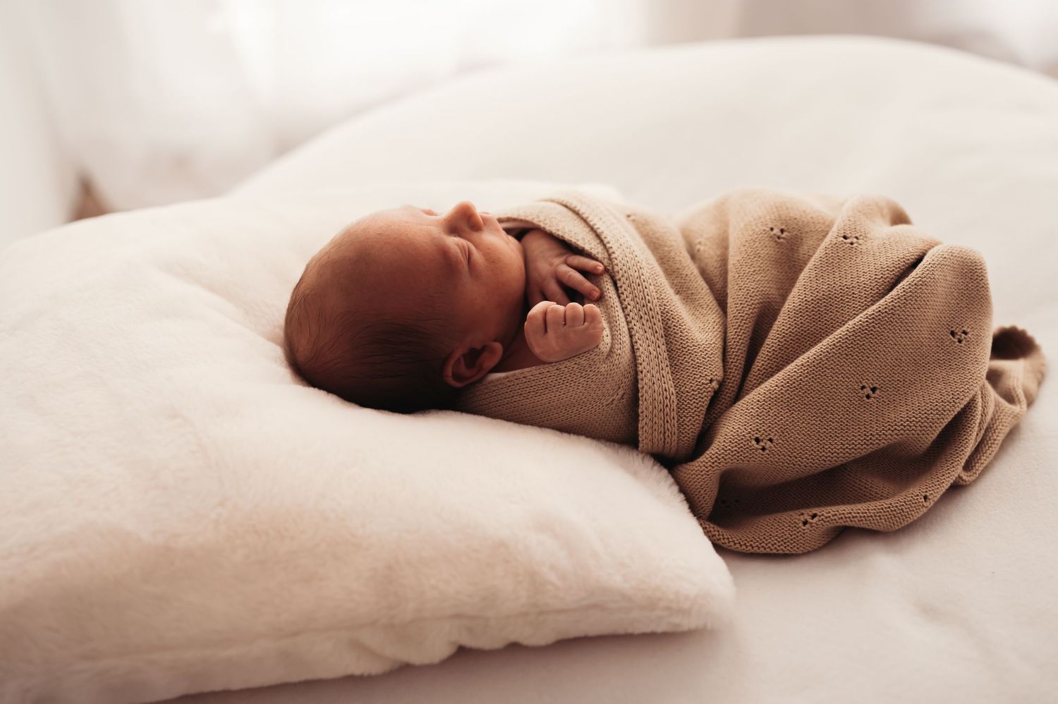 Baby yawning while swaddled in beige knitted blanket on white bedding.