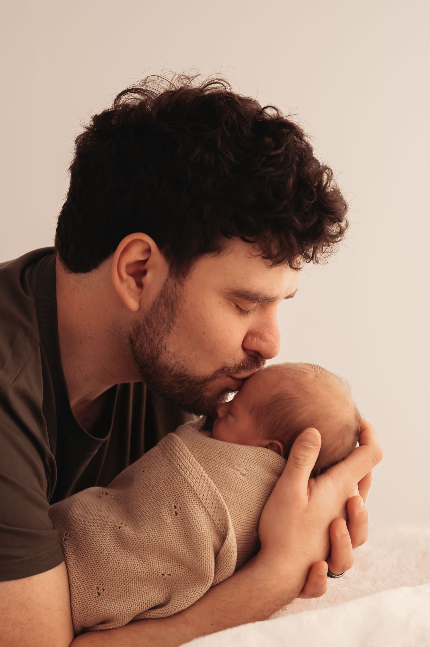 A person in a dark shirt gently kisses a swaddled infant's head against a light background.