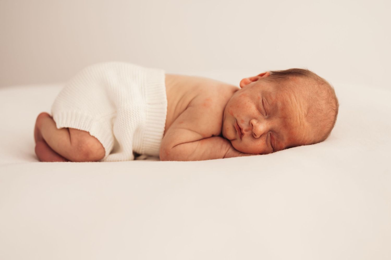 Sleeping newborn baby in white onesie lying on side against white background.