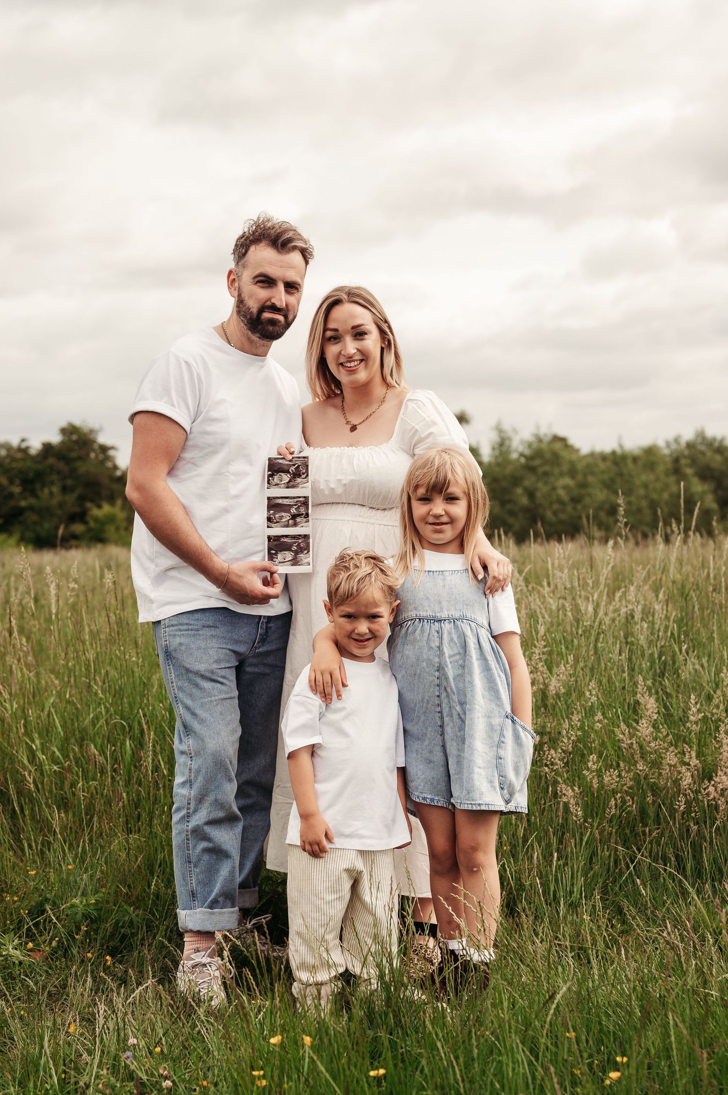 A family of four poses together in a grassy meadow while holding an ultrasound photo on a cloudy day.
