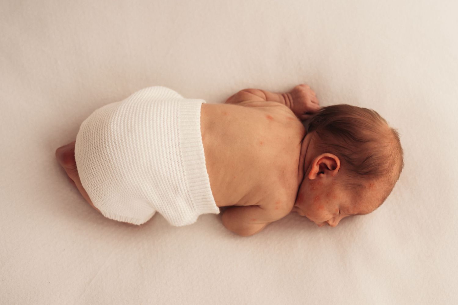 Newborn baby sleeping peacefully curled up on a white blanket, wearing white knit bloomers.