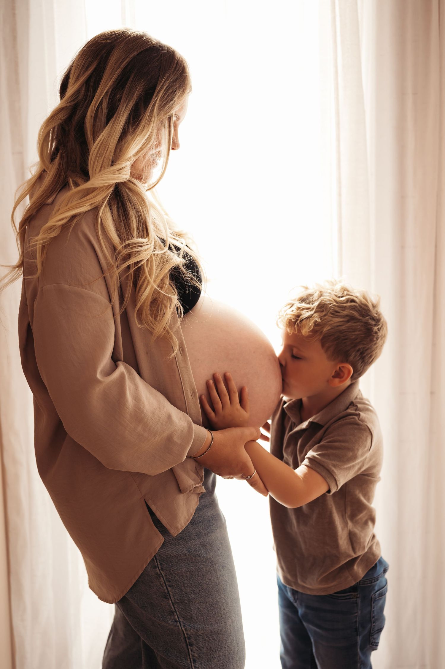 A young boy kissing his pregnant mother's bare belly while standing by a bright window in a tender moment.