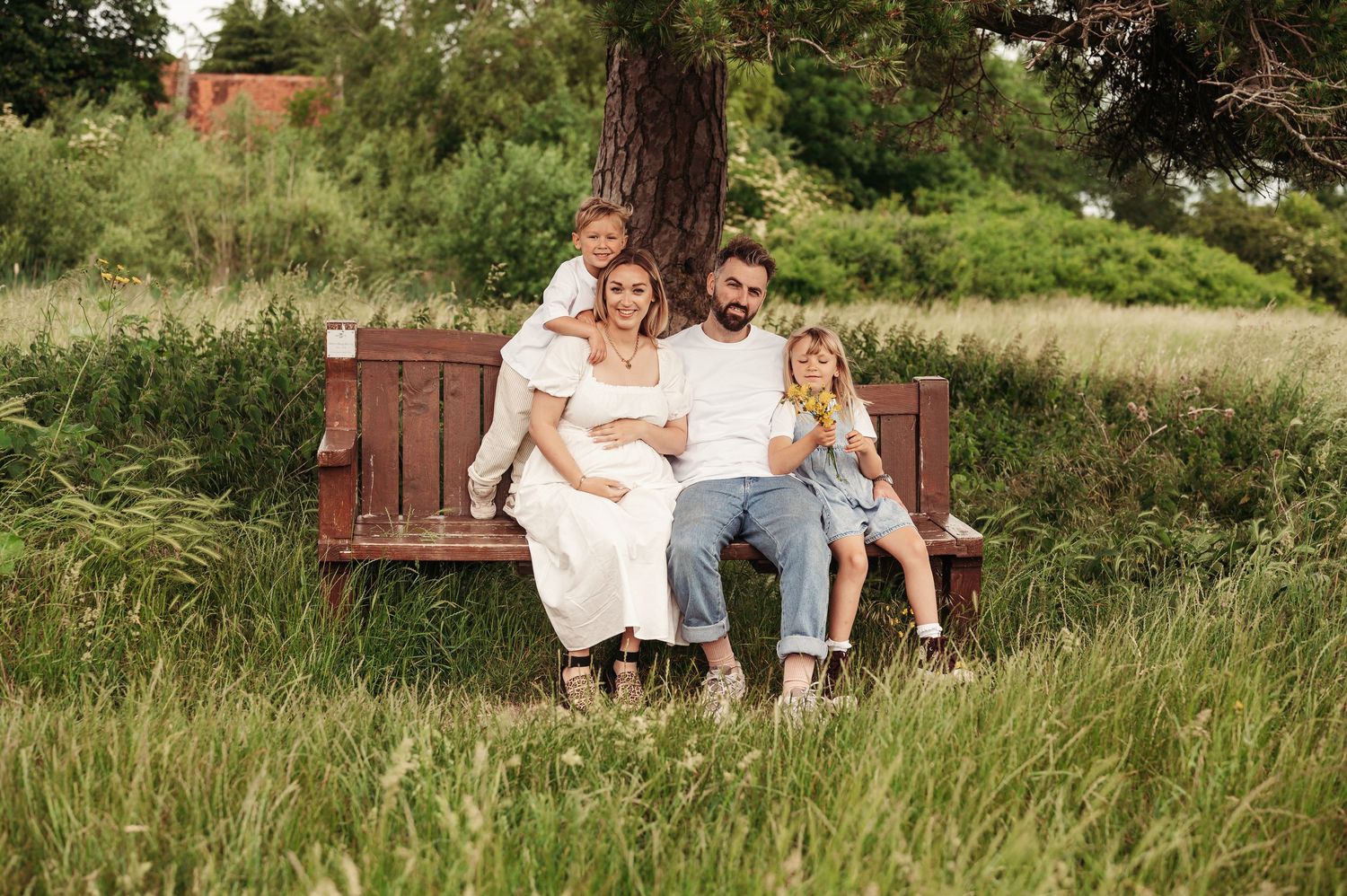 A couple sits together on a wooden bench surrounded by tall grass and greenery on a sunny day.