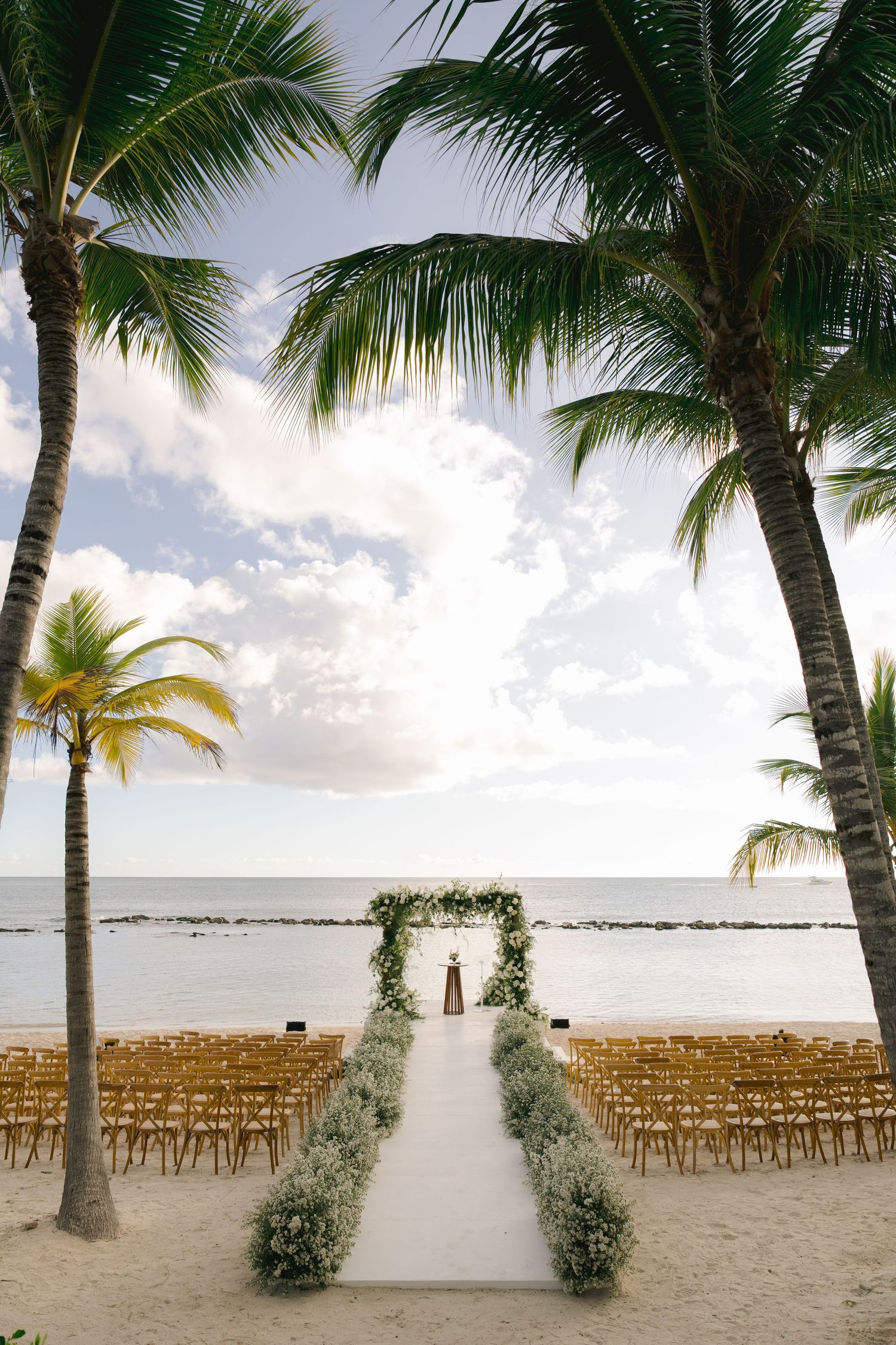 Beachfront wedding ceremony setup with wooden chairs and palm trees framing a white aisle runner on tropical sand.