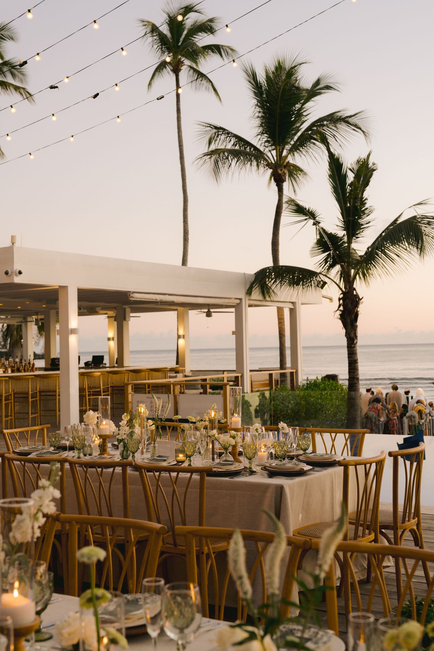 Elegant outdoor wedding reception setup with wooden tables and string lights under palm trees at sunset by the ocean.
