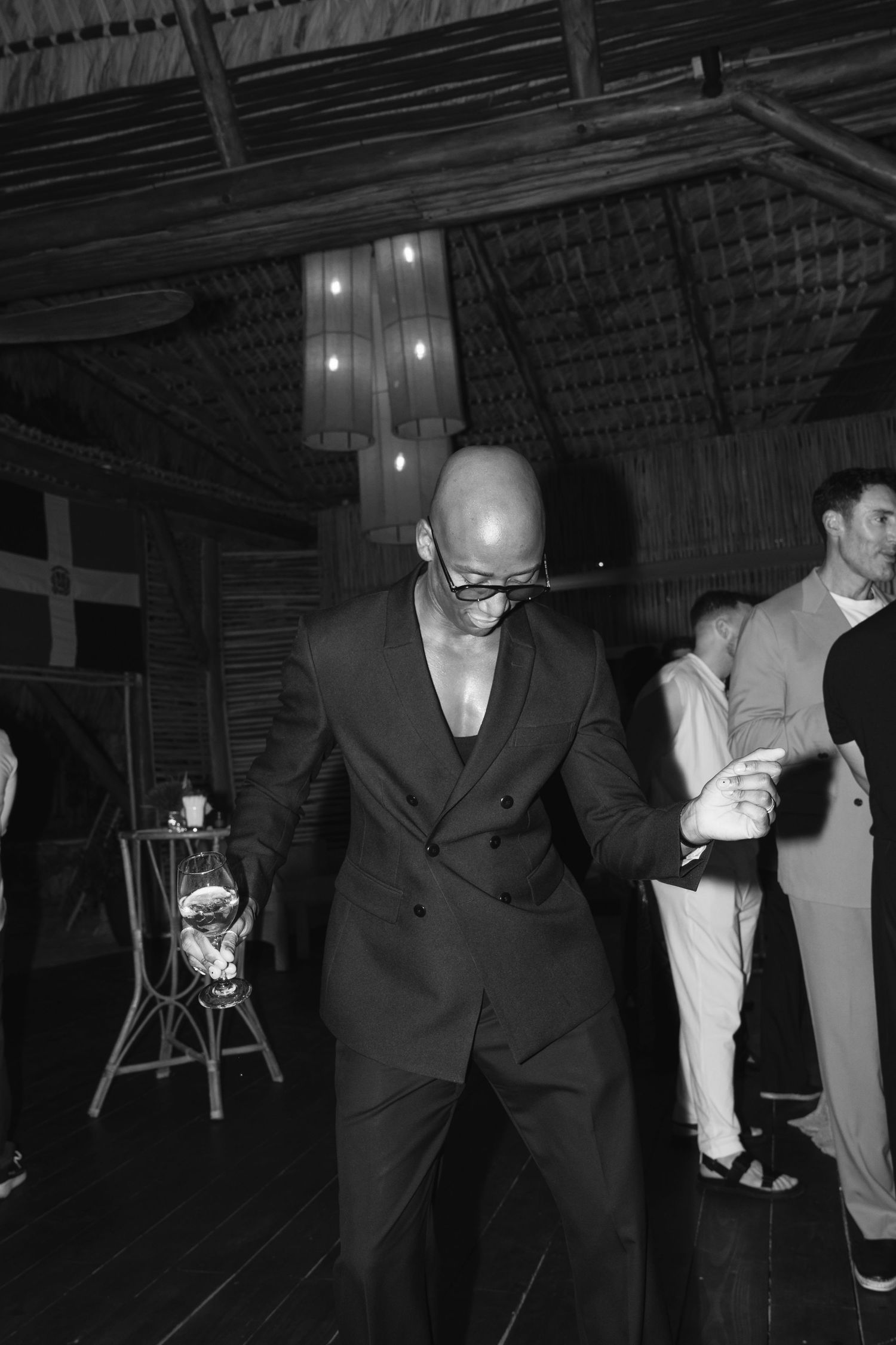 A person in a dark suit dances on a wooden floor under pendant lights in a rustic venue.