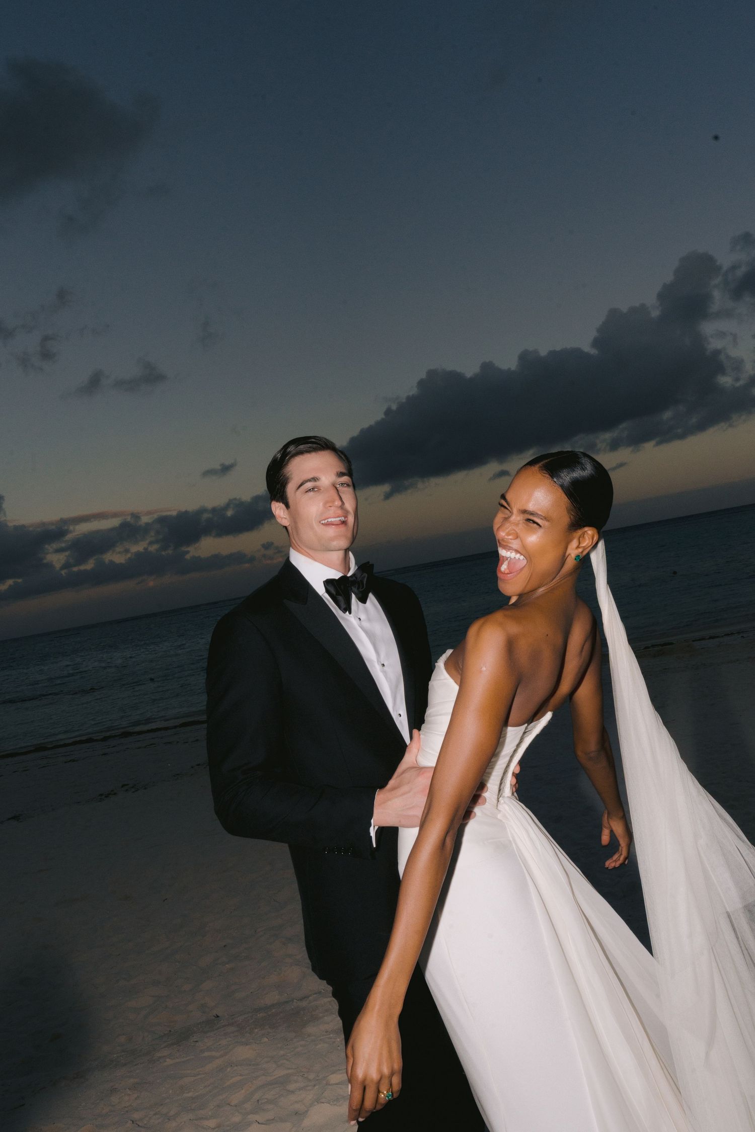 Newlywed couple celebrating on a beach at sunset, with the bride in a white gown and groom in black tuxedo.
