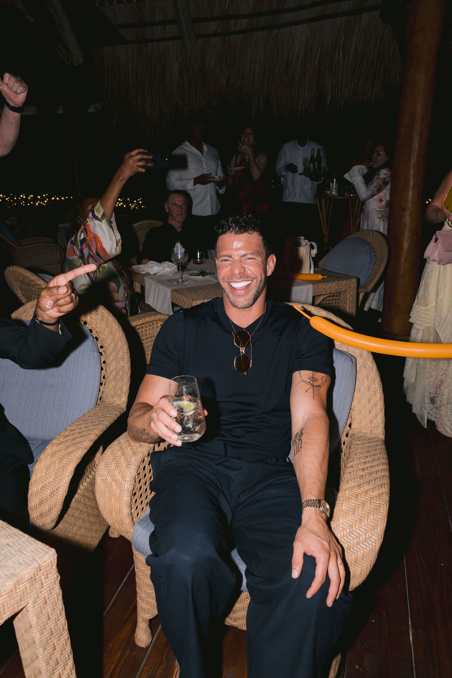 Someone in dark clothing sits in a woven rattan chair at a dimly lit venue while holding a drink.
