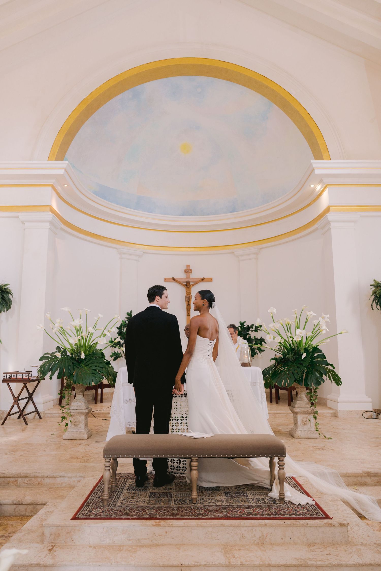 A couple stands at the altar in an elegant white chapel with a domed ceiling accented in gold trim and flowers.