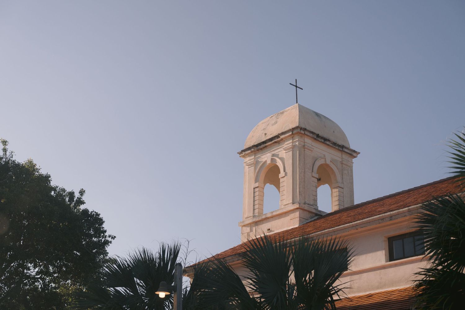 Church steeple with bell tower and cross rises above palm trees against a dusky blue sky.