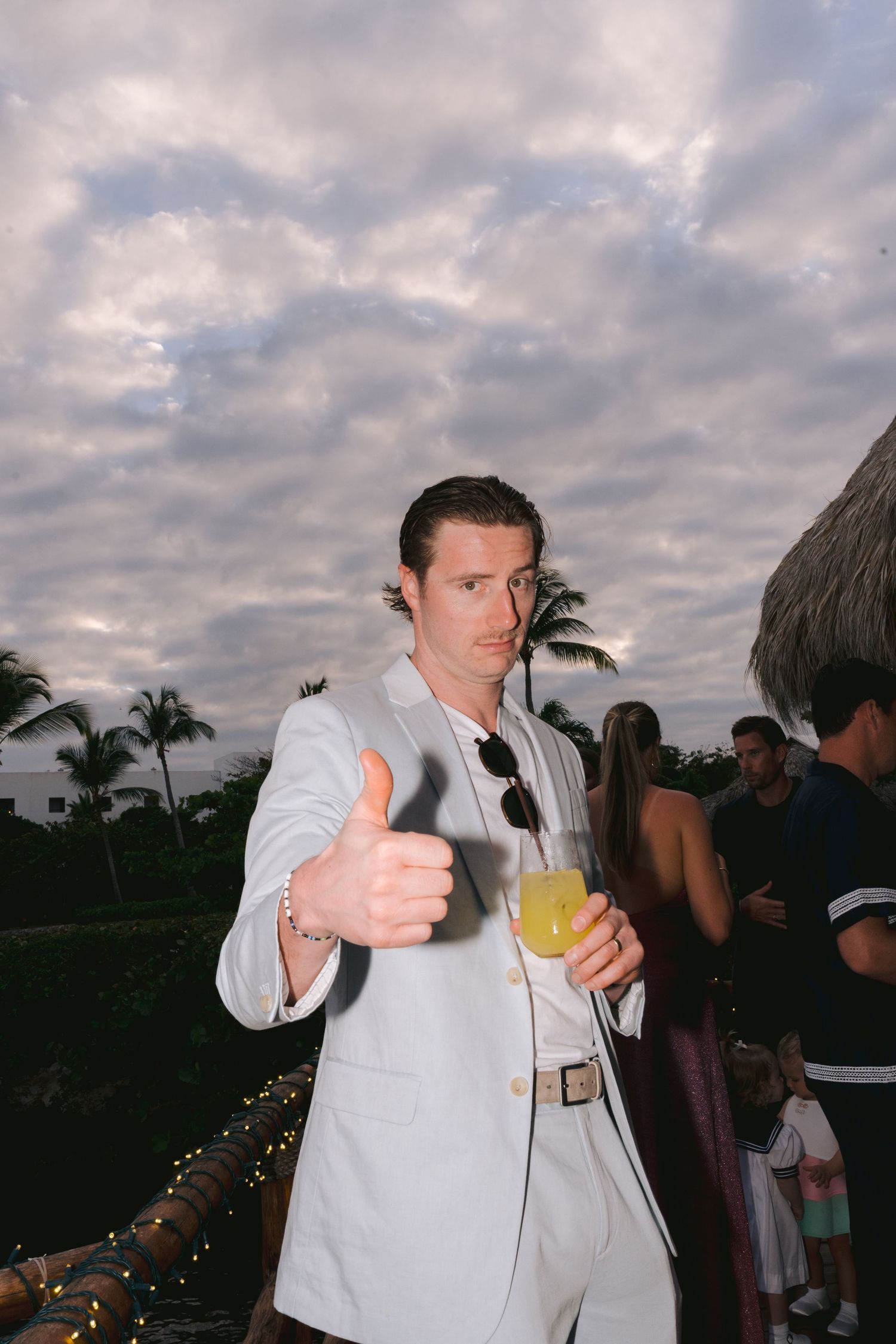 Someone in a light colored suit gives a thumbs up while holding a tropical drink at an outdoor sunset party with palm trees.