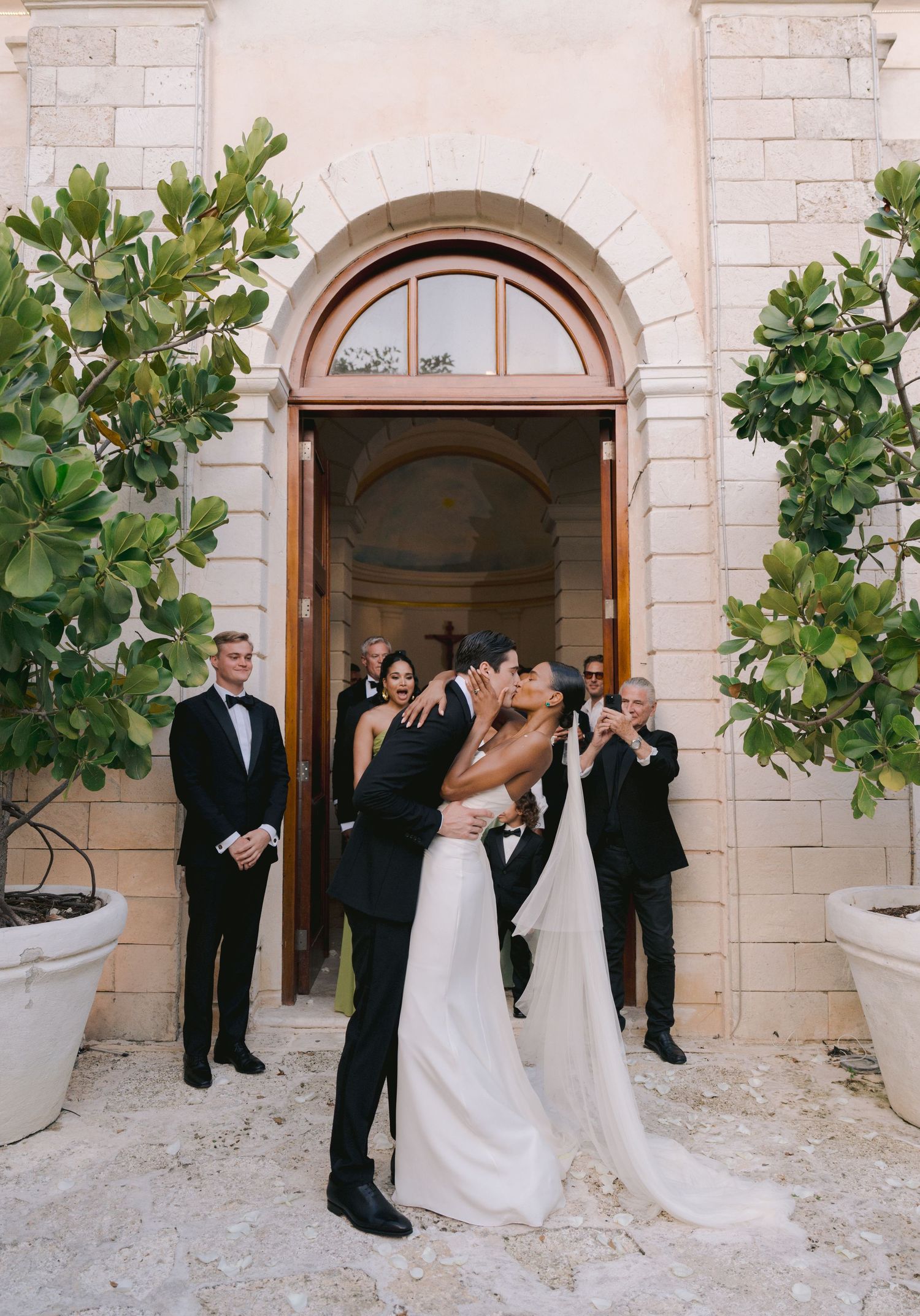 Wedding party gathered at arched entrance of limestone building with elegant green foliage framing the doorway.
