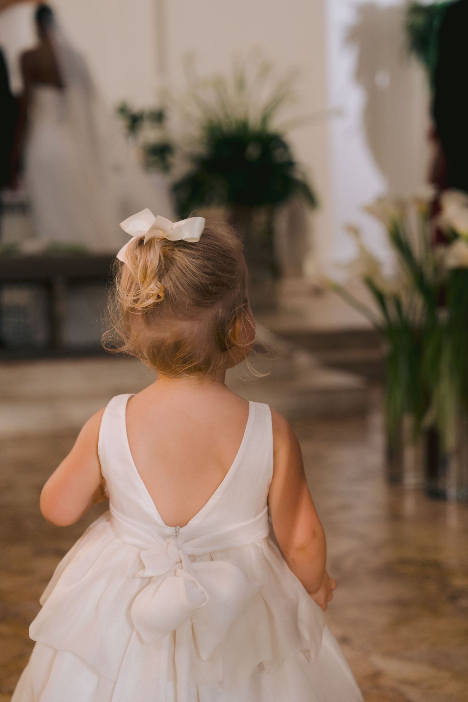 A toddler in a white formal dress with a bow stands in an elegant room with indoor plants in the background.