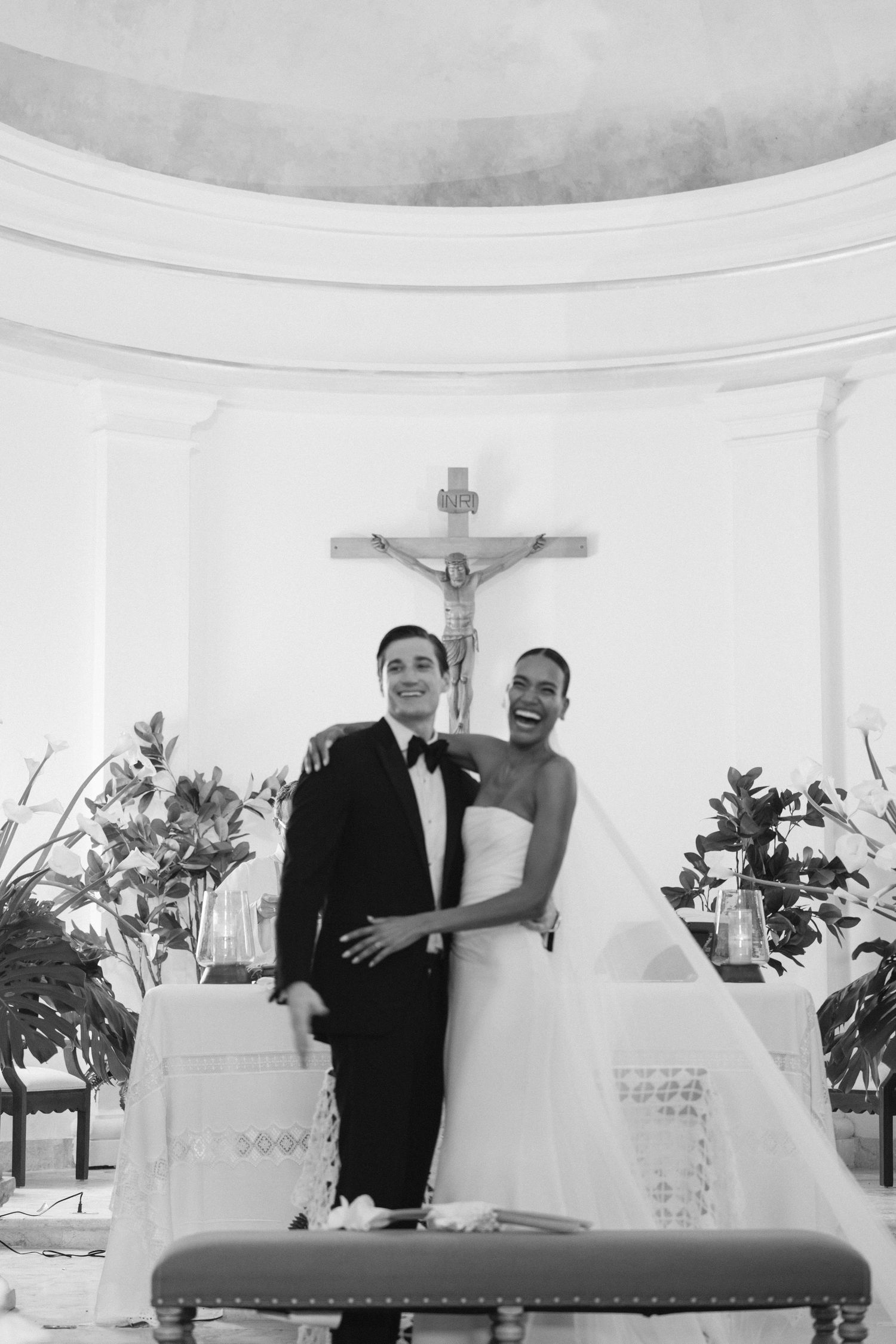 A joyful couple poses at the altar in front of a crucifix during their wedding ceremony in a classic church setting.