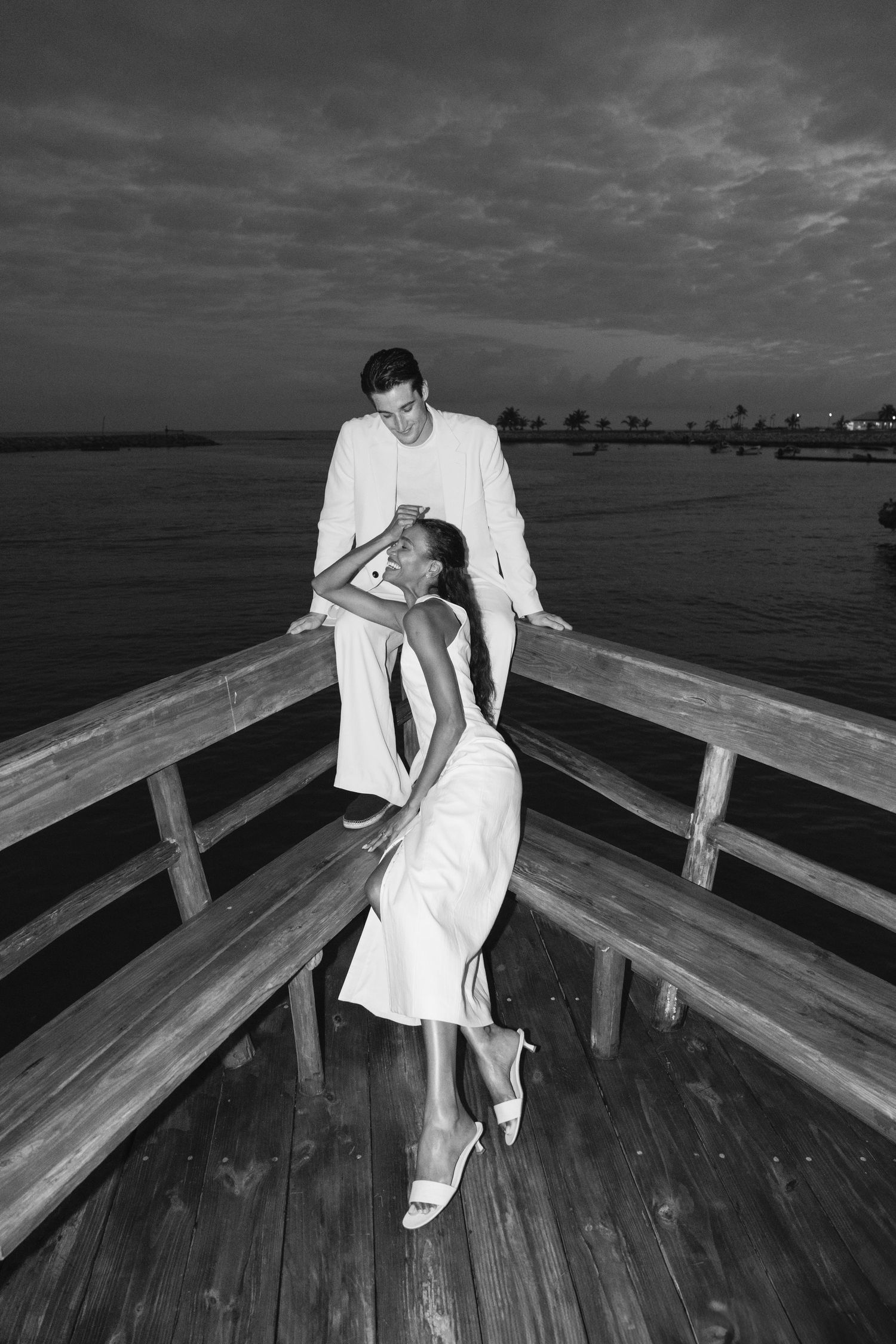 A romantic black and white photo of a couple in white outfits dancing on a wooden pier at dusk.