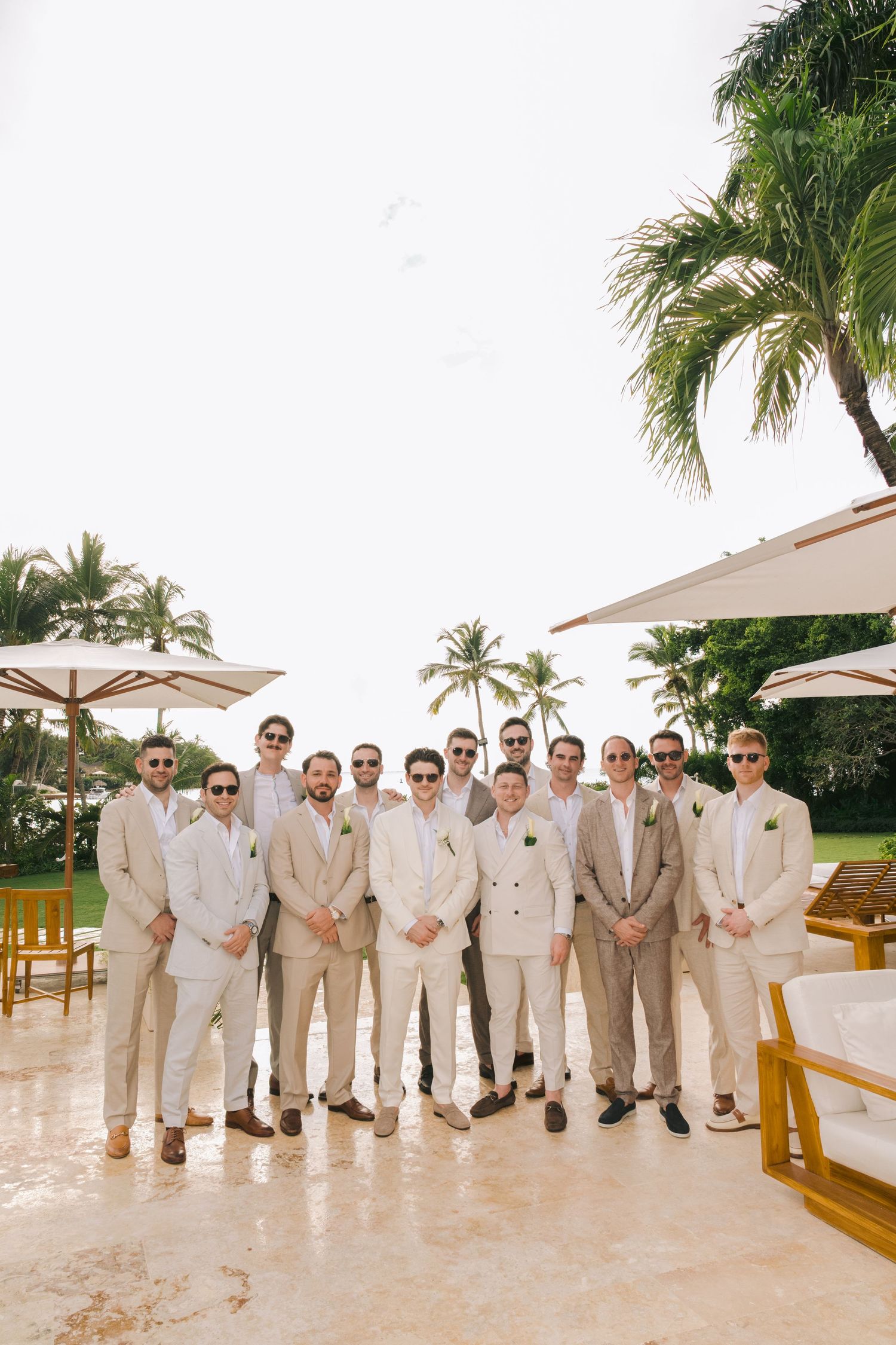 Wedding party in beige and tan suits pose together on a tropical resort patio with palm trees in the background.