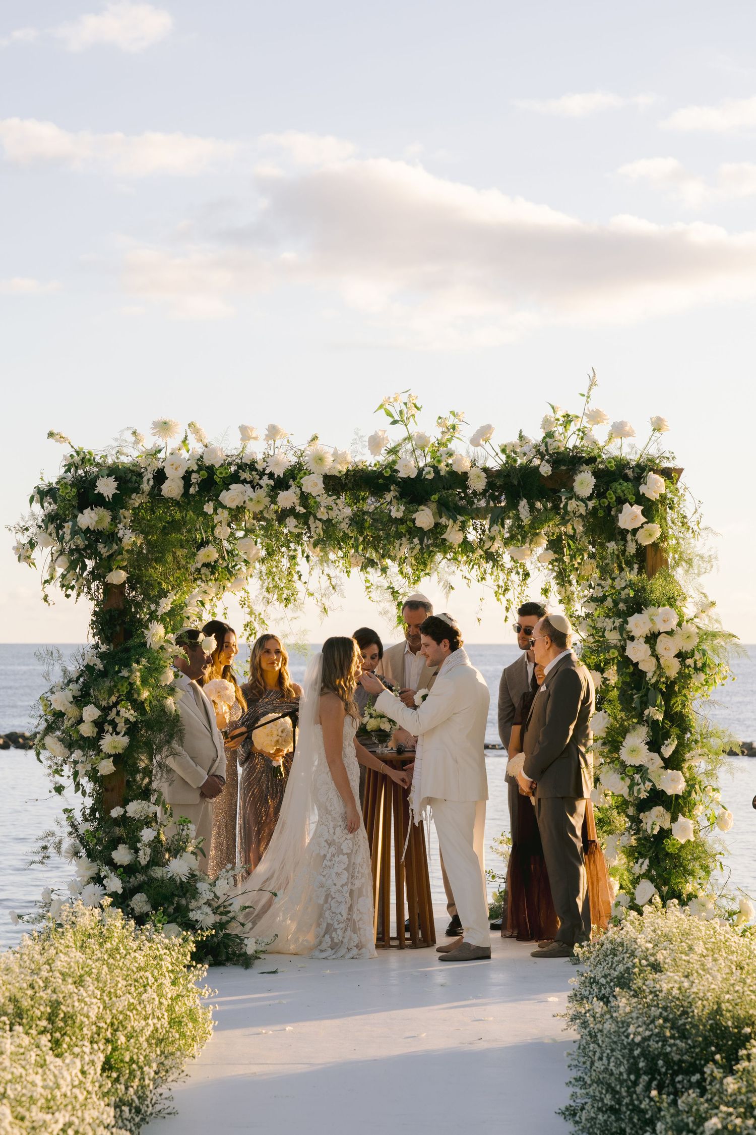 A beachside wedding ceremony takes place under a floral arch with white roses and greenery at sunset.