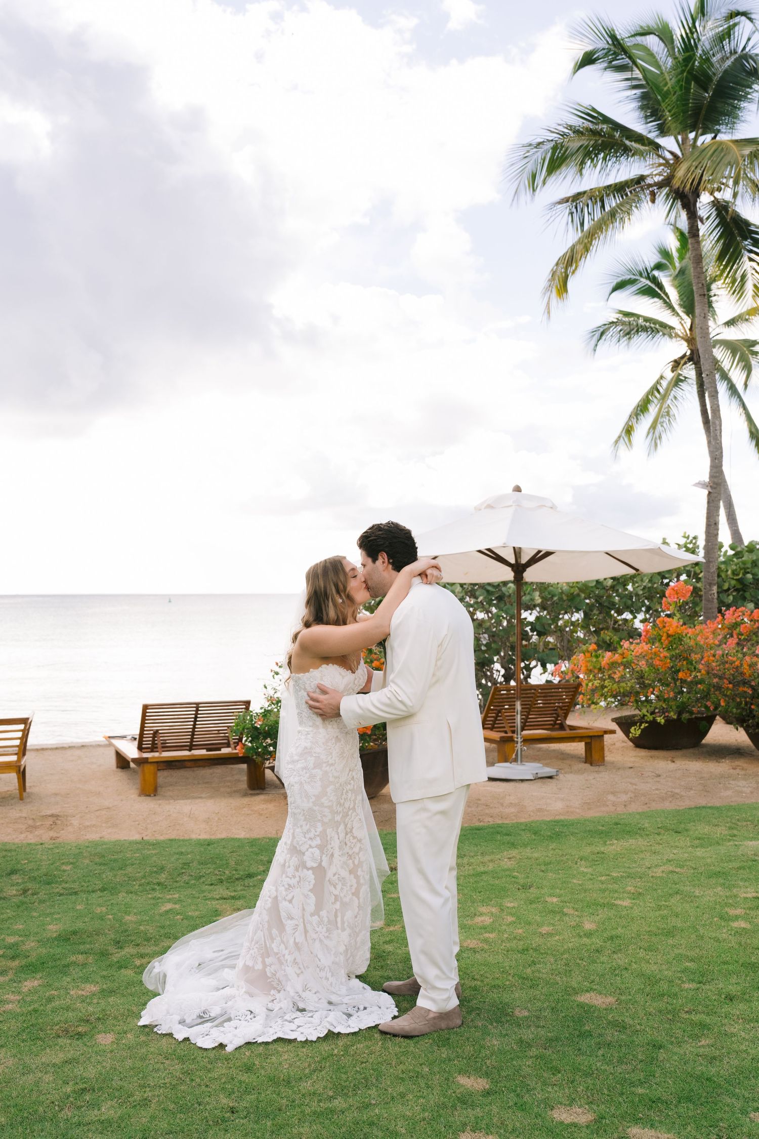 A romantic wedding couple shares intimate moments on a tropical beachfront lawn surrounded by palm trees and ocean views.