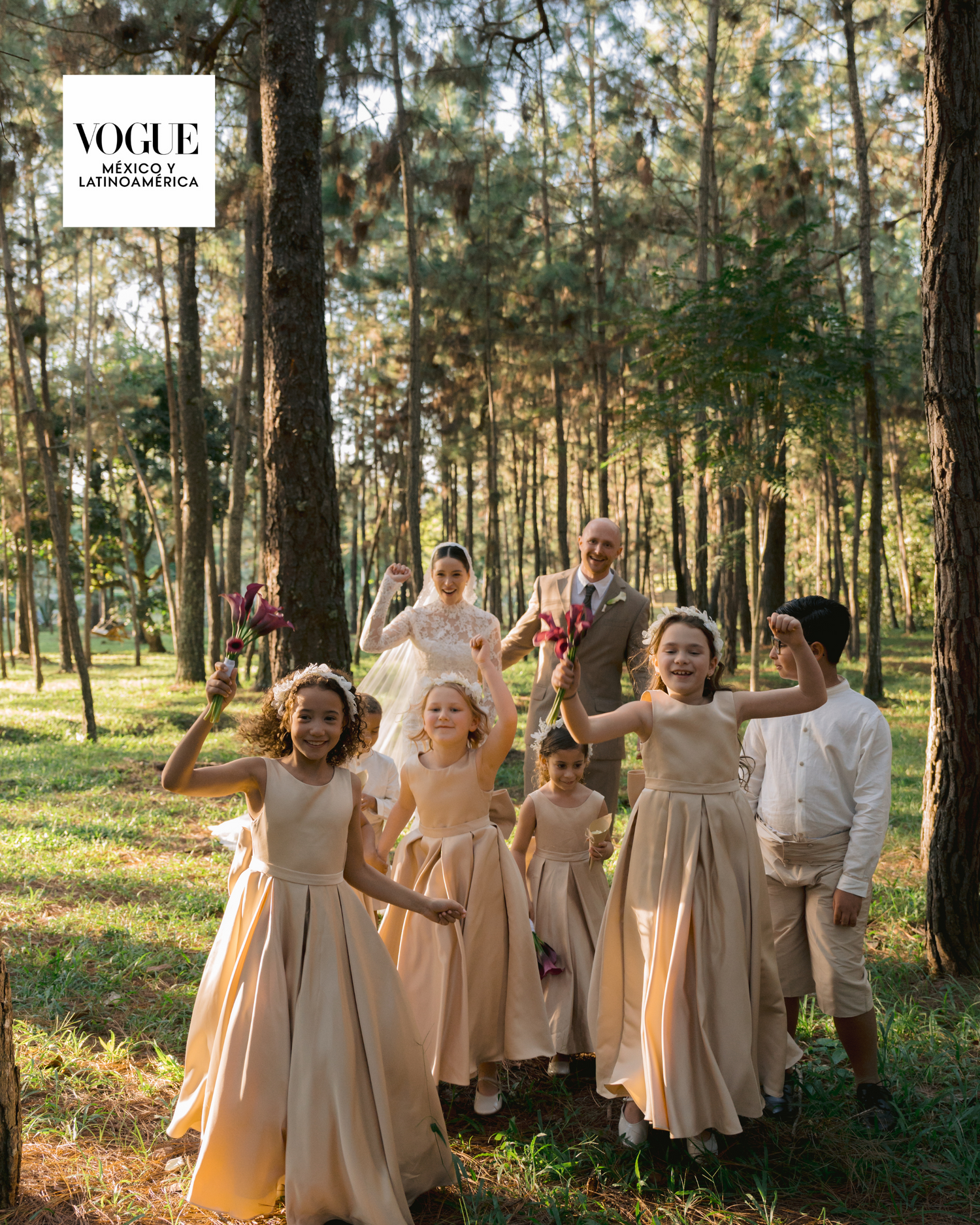 Wedding party in champagne-colored dresses poses together in a sunlit forest setting.