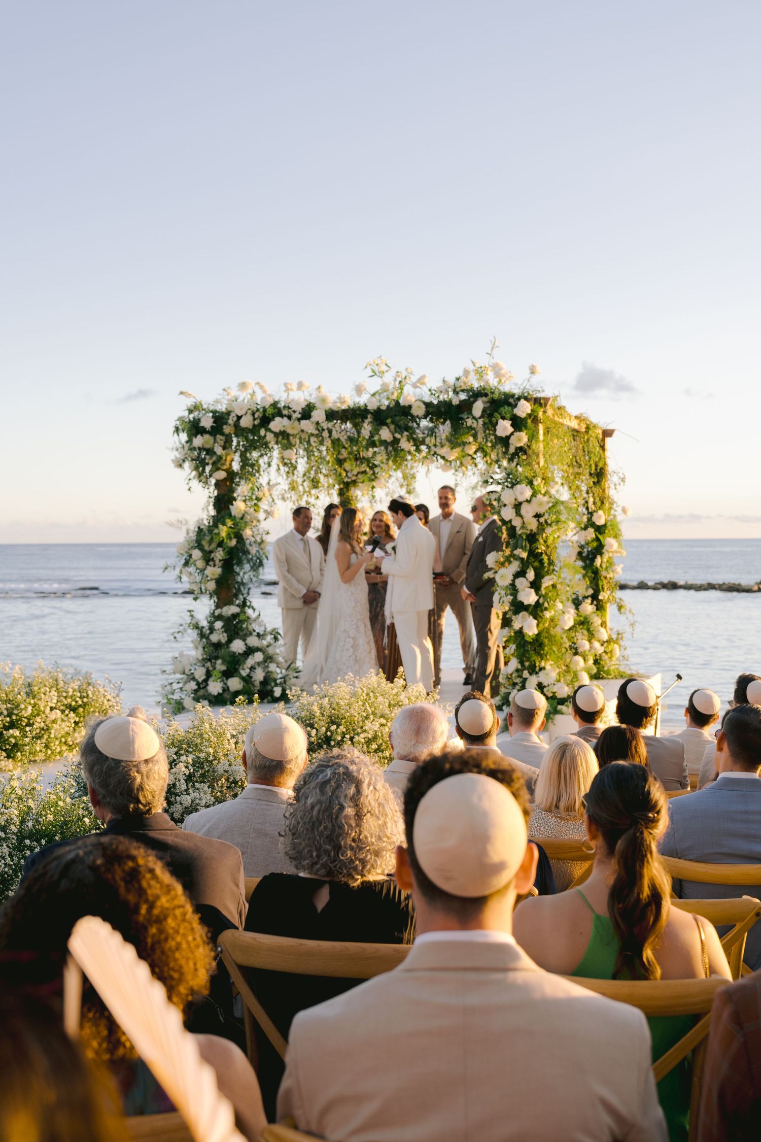 Guests watch an outdoor wedding ceremony under a floral arch by the ocean at sunset.