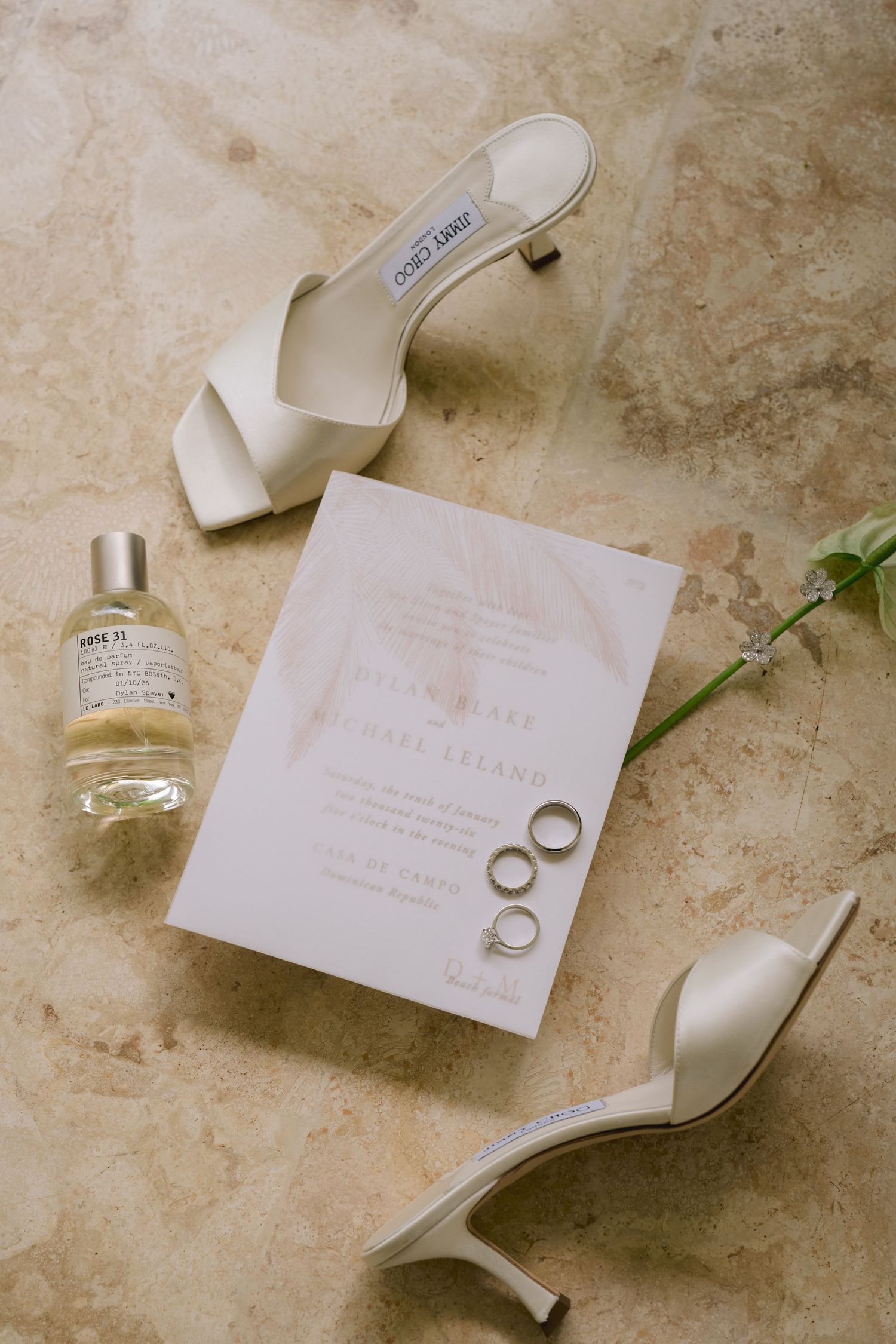 Wedding invitation and rings displayed elegantly with white heels on a textured beige stone surface.
