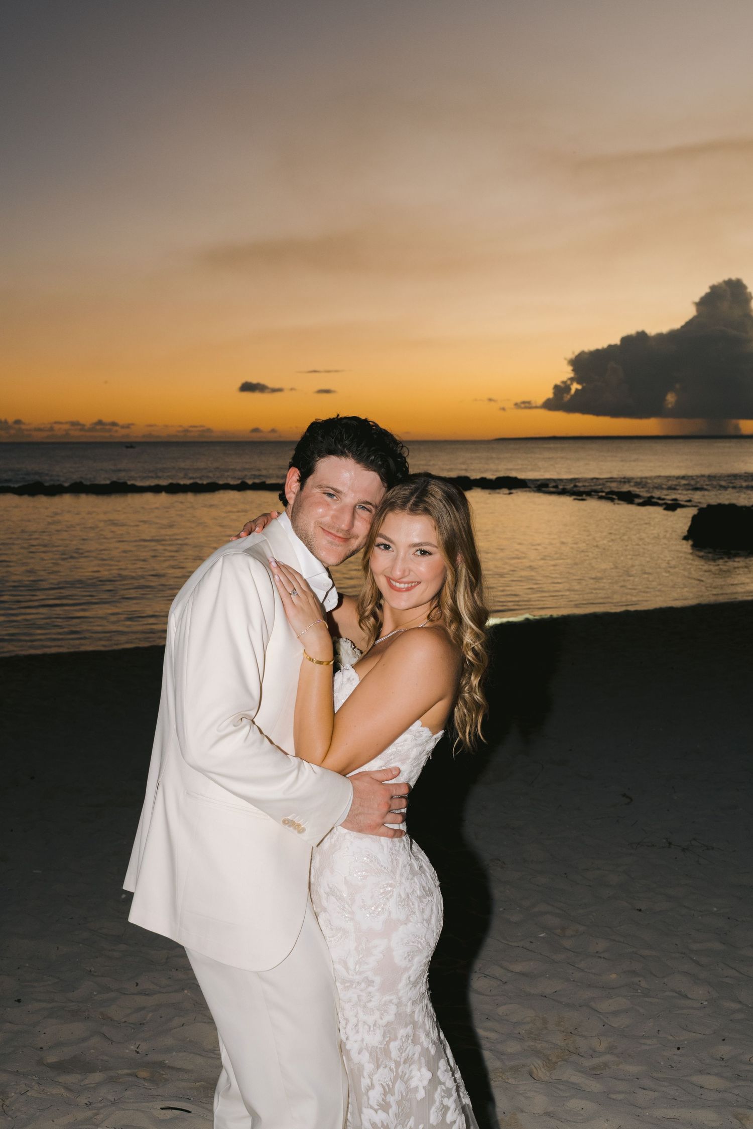 A couple embraces on a beach at sunset with golden orange sky and ocean waters in the background.